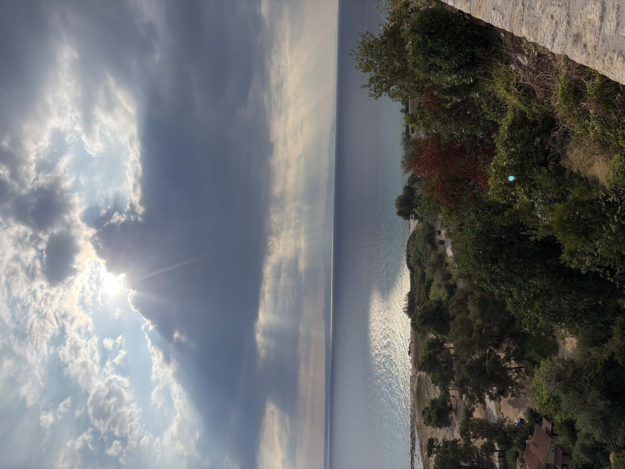 This is a landscape photograph taken from an elevated viewpoint overlooking the Aegean Sea. The scene captures a coastline with a sandy beach, dense green vegetation, and a body of water. Above, the sky is dramatic with a mix of dark, heavy clouds and patches of bright sunlight. Sunbeams are visible, breaking through the clouds and casting light onto the sea, creating a shimmering effect on the water's surface. In the distance, a faint outline of mountains or landmass can be seen across the sea. In the foreground, a stone wall runs along the right edge of the frame, and the lower portion of the image shows various trees and some buildings nestled amongst the foliage. The overall impression is one of a natural coastal setting, possibly during a break in stormy weather. The location is identified as being in the Kassandra Municipal Unit, Greece. There are no people or animals clearly visible in the image.