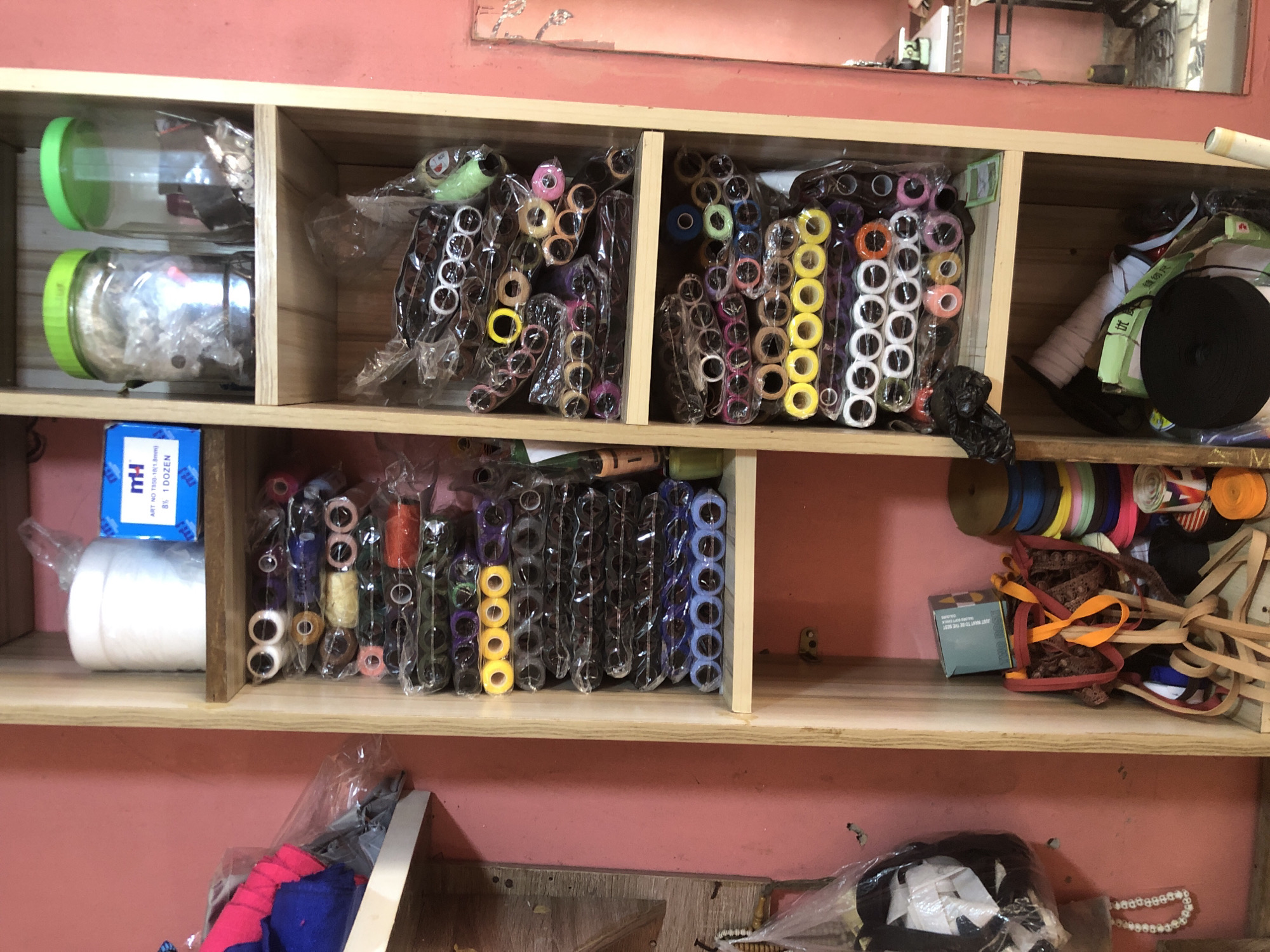 This image displays a well-organized wooden shelving unit filled with a variety of craft and sewing supplies, likely in a shop or workshop in Ado, Nigeria. The shelves are made of light-toned wood, set against a reddish-pink painted wall. Above the top shelf, a reflective surface, possibly a mirror, shows distorted reflections of the surrounding space.
On the top shelf, the left compartment holds two clear plastic jars with bright green lids, containing small items like buttons or beads. The central compartments are packed with numerous spools of thread, elastic, or ribbon in assorted colors such as yellow, pink, purple, white, orange, blue, green, and black. These are neatly arranged in clear plastic packaging. The top right compartment contains a large black spool of material, along with other items including a light green elastic and an object marked with the Chinese characters "纫尺" (meaning "sewing ruler").
The middle shelf features a blue and white box with visible text including "mTH", "ART NO T850-18(1.8mm)", and "8% 1 DOZEN", resting on a large white spool of thread or elastic. Adjacent compartments are filled with more colorful spools, similarly packaged. The right-hand section of this shelf is stacked with various rolls of colorful ribbons and elastic bands, alongside a small teal-colored box.
The bottom section of the image, visible below the shelves, shows fabric scraps in pink and blue contained within a clear plastic bag, and what appears to be a white beaded necklace or trim. No people are visible, and the scene depicts a static display of inventory, suggesting a retail or crafting environment.
