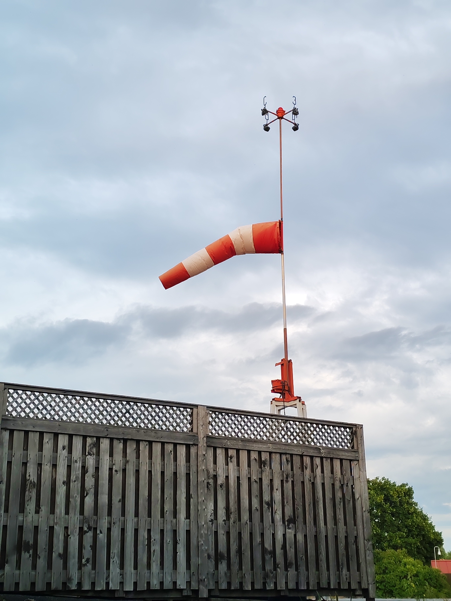 The image captures a windsock on a pole, a common sight at airports or other weather-sensitive locations. The windsock is orange and white, indicating wind direction and speed. Above the windsock, a weather instrument with rotating cups is visible. In the foreground, a wooden fence with a lattice pattern runs across the frame, partially obscuring the view of the windsock. The sky is overcast with clouds. The location is specified as Shalsky, Russia. There are no people or animals present in the image. The overall scene suggests an outdoor, possibly industrial or aviation-related setting.