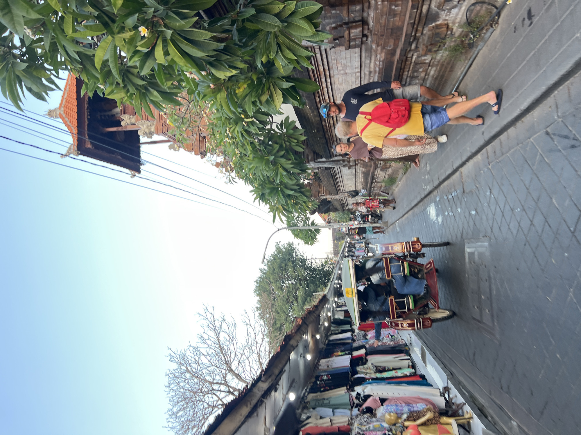 The image depicts a street scene in Kuta, Indonesia, likely an area with shops and traditional architecture. In the foreground, a red and gold pedicab, or "becak," is traveling down the street, carrying passengers. The pedicab has a roof and appears to be made of wood and metal. To the left of the street, a row of market stalls is visible, with clothing and other goods displayed on racks. The stalls have awnings to provide shade.
Further down the street, several people are walking. One person in the foreground is wearing a yellow t-shirt and blue shorts, with a red backpack. Another person, a man in a dark t-shirt and shorts, walks beside him. A woman in a brown top and patterned skirt is also walking in the same direction. In the background, more people and motorbikes are visible, suggesting a bustling urban environment.
To the right of the street, a wall constructed of reddish-brown bricks is prominent. Above the wall, lush green foliage from trees with white flowers is abundant, with a traditional Balinese-style building with an orange-tiled roof visible behind the trees. The sky above is a clear, pale blue, indicating it is likely daytime, possibly late afternoon or early morning due to the soft lighting. The street itself is paved with interlocking stone pavers. The overall impression is a vibrant street market in a tropical location.