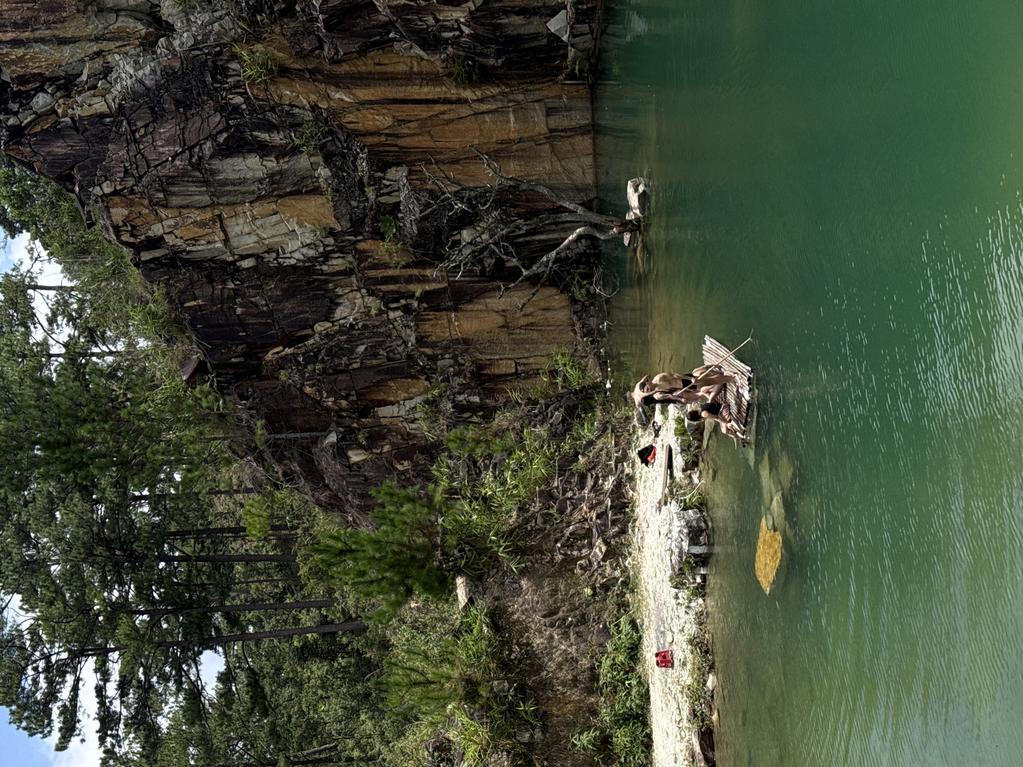 The image depicts a quarry lake in Lán Tranh, Vietnam. A group of people are on a bamboo raft in the emerald green water. The raft is positioned in the foreground to the left of a large, imposing rock face that dominates the background. The rock face is striated with various colors, including brown, tan, and grey, indicating its geological composition. Lush green vegetation, including pine trees, grows along the top of the quarry and on the slopes leading down to the water. The water itself is clear and calm, with visible ripples on the surface. On the shore to the left, there is a red object, possibly a life vest, and some bags. The overall scene suggests a recreational activity in a natural, rugged environment.