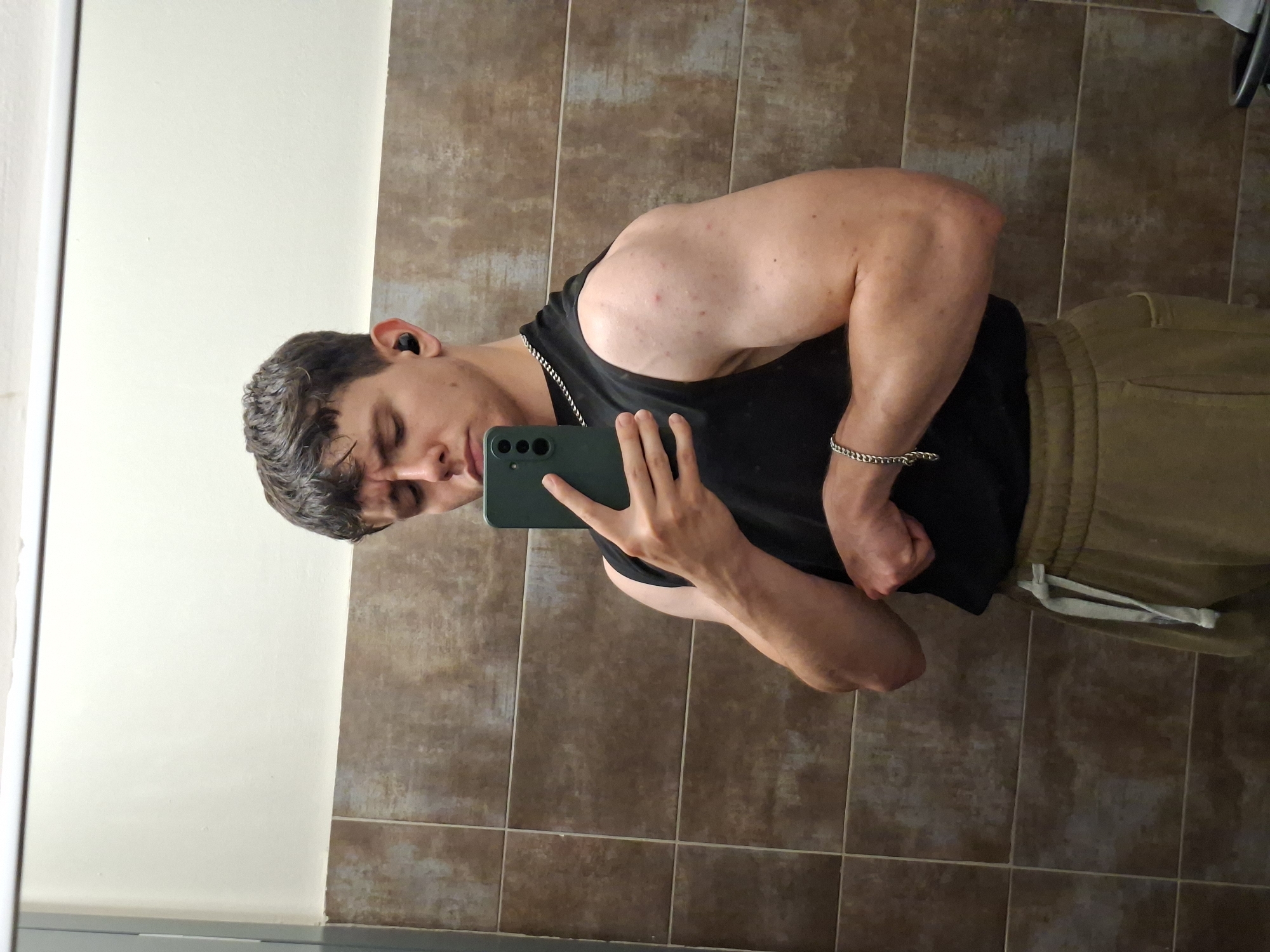 The scene depicts a young adult male capturing his reflection in a mirror within an interior space in Plovdiv, Bulgaria. The male, appearing muscular, wears a black sleeveless top, olive-green or khaki sweatpants with a light drawstring, a silver-toned chain necklace, and a matching bracelet on his right wrist. A black wireless earbud is visible in his left ear. He holds a dark green smartphone, featuring a triple camera array, in his left hand, positioned for a selfie. His right arm is flexed, showcasing his bicep. His hair is dark, short, and slightly textured. Visible reddish blemishes are present on his right shoulder and upper arm. The background consists of a lower wall section tiled with rectangular, brownish-grey tiles, and an upper section of plain, light off-white or cream-colored painted wall, separated by a horizontal trim. A light-colored vertical partition or frame is visible on the far left.