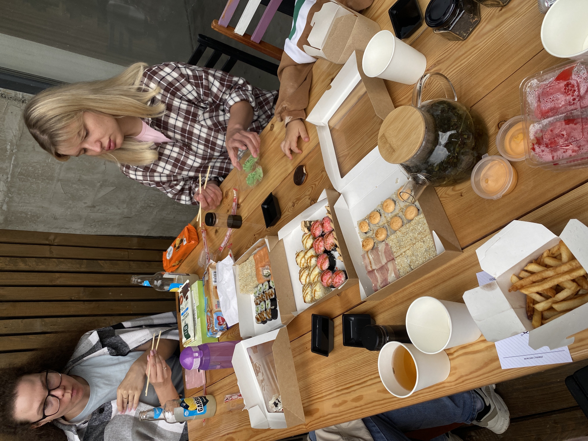 The image shows a group of people, likely a family or friends, sharing a meal outdoors at a wooden table. The setting appears to be a casual dining area, possibly a cafe or a restaurant patio, in the city of Фоки, Russia. The table is laden with various food items, including sushi, french fries, and what appears to be tea in a glass teapot. There are also drinks in bottles and cups, as well as small containers of sauces and what might be wasabi. Two women are visible, one with curly hair and glasses, wearing a plaid shawl, and another with blonde hair wearing a plaid shirt. A child's hand is also visible reaching for food. The overall atmosphere suggests a relaxed and enjoyable gathering.