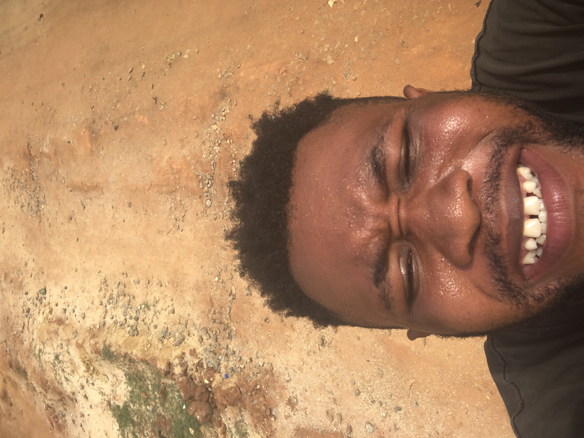 The image displays a close-up of a person's head and upper torso, lying on sandy, reddish-brown ground. The individual, who appears to be a man with dark skin, short curly hair, and a beard or stubble, is captured in a moment of intense expression. His eyes are tightly closed, forehead and nose crinkled, and mouth wide open in what appears to be hearty laughter or a strong emotional reaction, revealing white teeth and pink gums/tongue. A visible sheen of sweat covers his forehead and face, suggesting warm weather or exertion. He is wearing a black shirt.
The setting is an outdoor area with uneven, light reddish-brown earth. The ground is characterized by a mix of fine sand, scattered small pebbles, larger stones, and some patches of dry, sparse vegetation. The texture varies across the surface, with some areas appearing smoother and others more rugged. The lighting is bright, indicating it is daytime. No text is visible in the image.