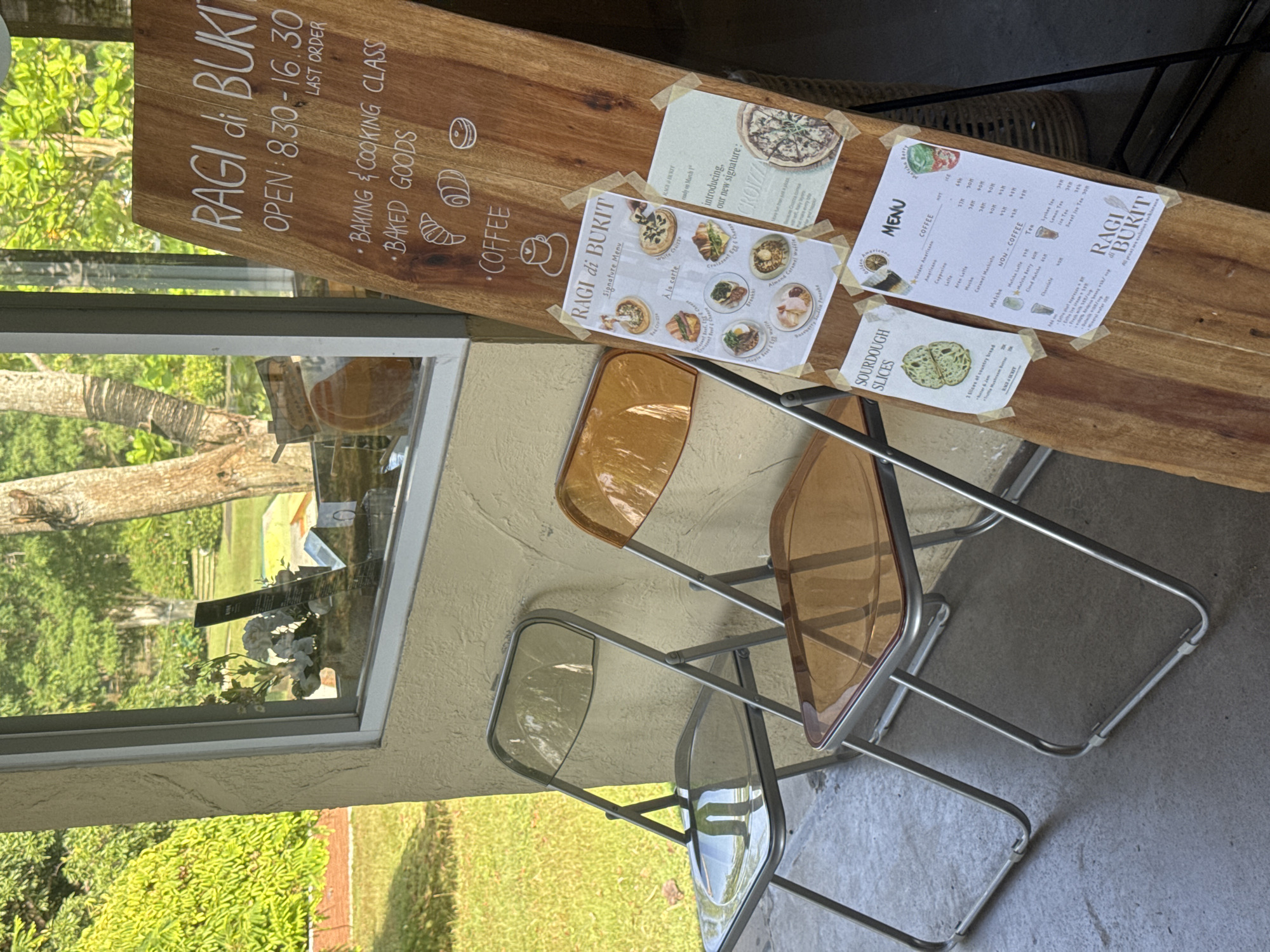 Outside Ragi di Bukit in Kuta Selatan, Indonesia, a wooden sign displays the cafe's name, hours (8:30-16:30), and offerings: baking classes, baked goods, and coffee. Menu cards taped to the sign feature signature dishes, sourdough slices, coffee, and non-coffee beverages. Two transparent chairs (one amber, one clear) sit in front of the sign. A window reflects trees and sky.