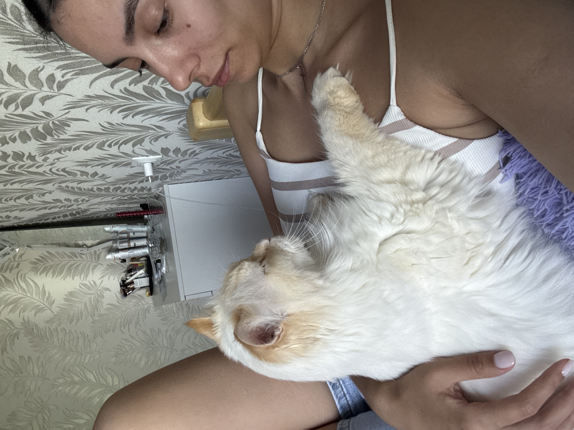 A woman is holding a white cat with light orange markings. The woman wears a white tank top and denim shorts. The cat is resting against her chest. The background includes a patterned wallpaper and a vanity with various items on it. Based on the prompt, the location is Chaikovsky, Russia.