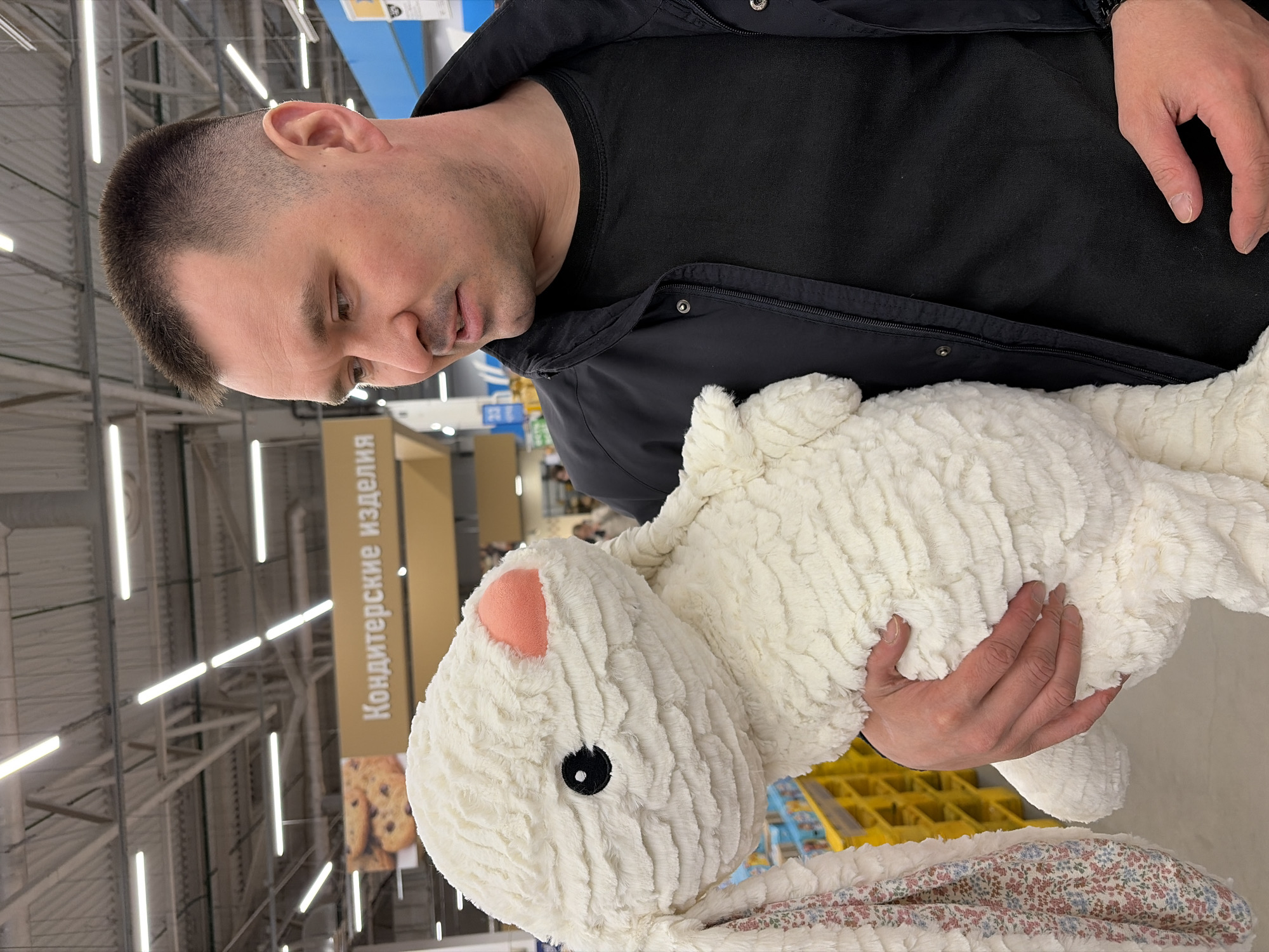 A man stands inside a store in Izhevsk, Russia, holding a stuffed white bunny. He wears a black shirt and jacket. A sign in Russian, "Кондитерские изделия" (Confectionery), is visible behind him. The store has fluorescent lights.