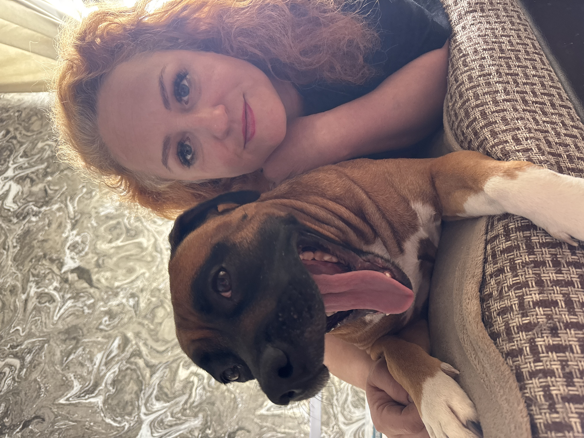 The image presents an upward-angled, close-up shot of a woman and a dog resting together indoors. The woman, positioned at the top of the frame, has reddish-brown, curly hair and fair skin, with a gentle smile and blue eyes looking directly at the viewer. She is wearing a dark top, and her arm is visible around the dog, suggesting a close bond.
Below the woman, a medium-sized dog, resembling a Boxer with a brown coat, black muzzle, and white markings on its chest and paws, is visible. Its tongue is hanging out, indicating relaxation or panting, and it appears to be resting comfortably. The dog is nestled in what looks like a dog bed or cushion, characterized by a woven brown and beige geometric pattern.
The background primarily features a grey and white abstract or marble-like swirled pattern. A soft light source is faintly visible in the upper left portion of the image. The overall impression is one of affection, peace, and contentment shared between the woman and her pet within a cozy indoor environment. No text is visible in the image. This scene is contextualized as being in Chaikovsky, Russia.