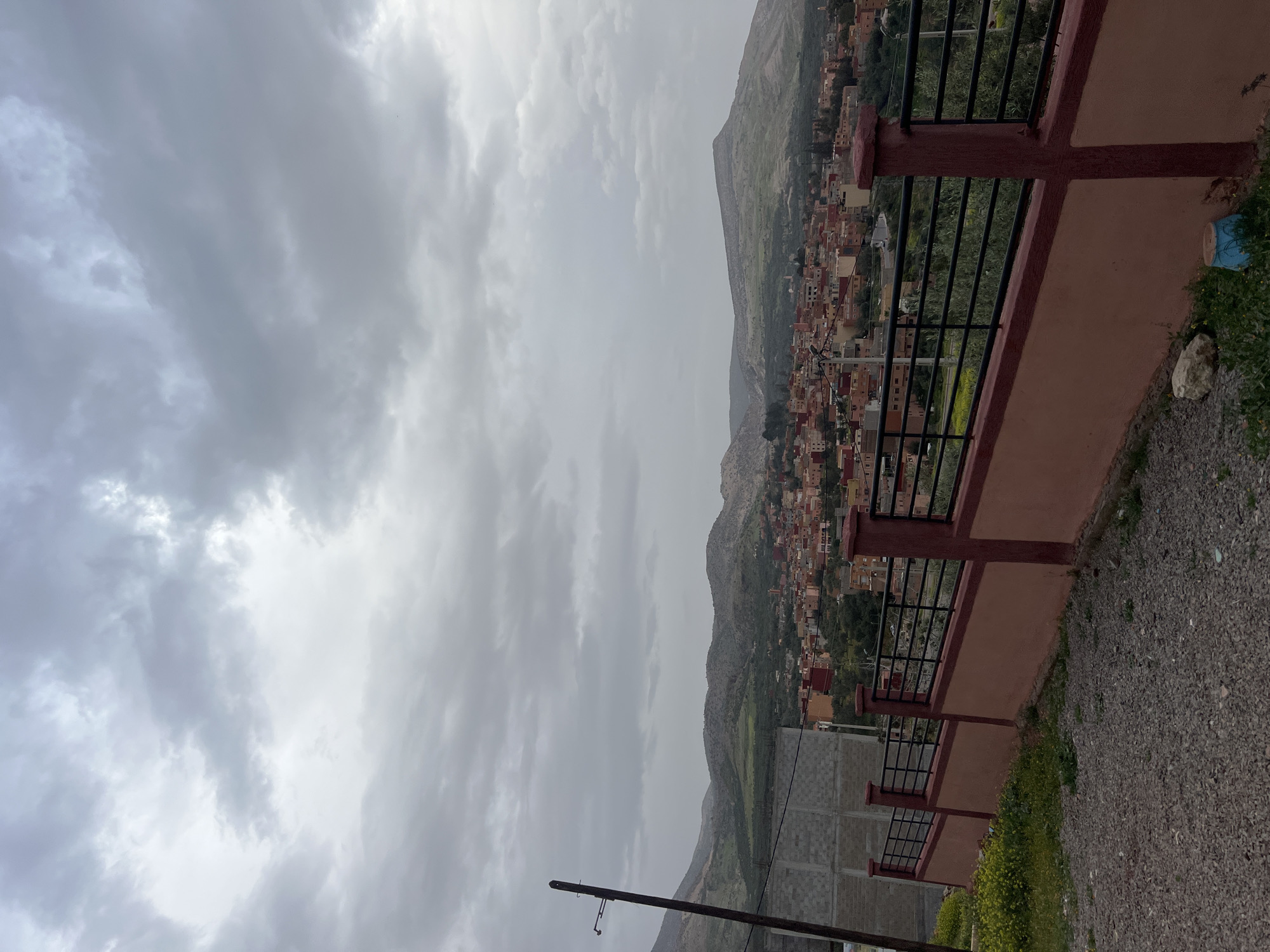 The image shows a view from what appears to be a residential area overlooking the town of Ouaouizert, Morocco. The sky is overcast with grey clouds. A mountain range is visible in the background behind the town, which consists of tightly packed buildings. A reddish-brown concrete wall with black metal railings lines the right side of the frame, likely part of a balcony or terrace. The ground near the wall is covered with gravel and sparse vegetation. In the lower left corner, a power pole with wires is visible. The overall scene suggests a cloudy day in a mountainous region with dense urban development.