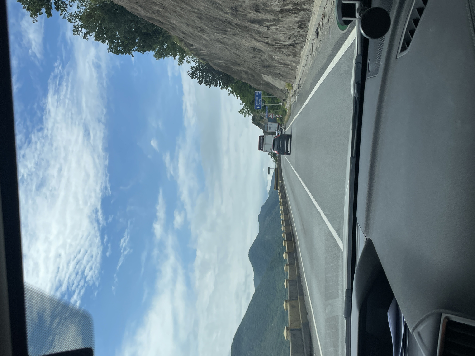 The image displays a daytime view from inside a vehicle traveling on a highway through a mountainous region. The perspective is looking forward through the windshield, with parts of the dashboard visible in the foreground on the right, including vents and a mounted device.
The highway features at least two lanes in the direction of travel and appears well-maintained. On the left side of the road, a steep, rocky cliff face is covered with lush green trees. To the right, a mountain range extends into the distance, with a concrete guardrail lining the highway's edge. The sky is bright blue, dotted with scattered white, wispy clouds.
Several vehicles are visible ahead on the highway, including a large white truck followed by a dark-colored SUV or car. A prominent blue road sign on the left of the highway provides directional information, clearly stating: "↑ Turnu-Severin 10km" and "Orșova." Below these, the text "Portile de Fier I" (Iron Gates I) is partially visible. These location cues, combined with the context, indicate the scene is in Romania, near Drobeta-Turnu Severin.