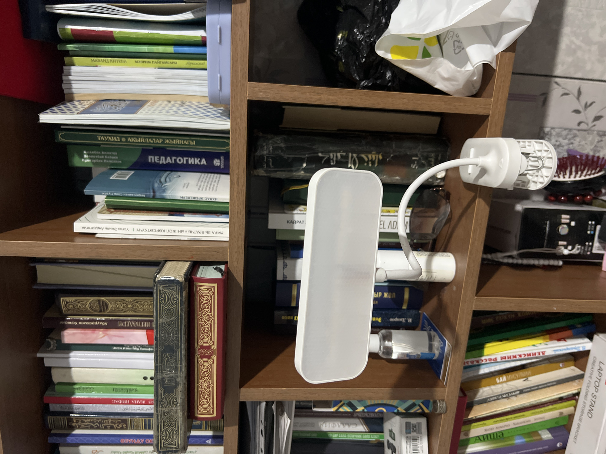 The image shows a bookshelf filled with books. The books are arranged in rows and columns on multiple shelves. Some of the books are thick and appear to be older, while others are thinner and more modern. There are also some other objects on the shelves, including a small bottle of liquid, a white clip-on lamp, and a plastic bag. The bookshelf is made of wood and appears to be old. The room is dimly lit, and there is a shadow cast on the wall behind the bookshelf. The image is taken from a slightly elevated angle, looking down at the bookshelf. The overall impression is one of a cluttered but organized space.