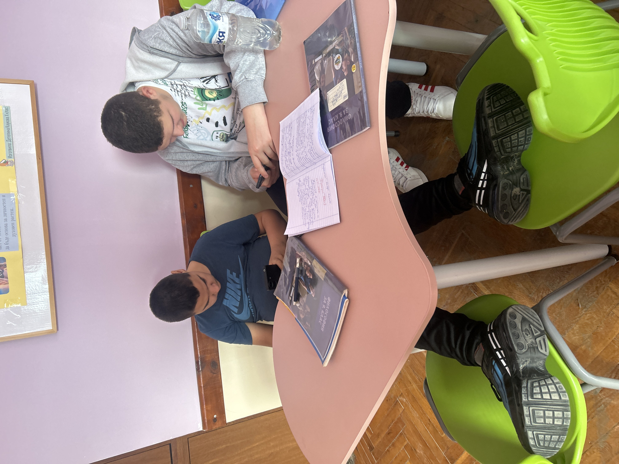 Two young individuals are seated at a light pink table in what appears to be a classroom or study area, suggested by the bright lime green chairs and instructional materials. The setting features a light purple wall and a wooden parquet floor, indicative of an indoor environment in Zlatograd, Bulgaria.
The subject on the left, wearing a gray hooded sweatshirt over a white graphic T-shirt, is actively engaged with an open notebook, holding a pen, and seemingly writing or studying its contents. A clear plastic water bottle with a partially visible "КЯ" label rests beside his right arm. He also wears white sneakers with red stripes, partially visible under the table.
The subject on the right, clad in a dark blue T-shirt with the "NIKE" logo prominently displayed, is looking down, possibly at a smartphone which is partially visible on the table near him, or at a book. His feet, wearing black and blue athletic shoes, are seen resting on the base of his chair.
On the table, besides the notebook, are two textbooks, both clearly titled "ФИЛОСОФИЯ ЗА 9. КЛАС" (Philosophy for 9th Grade), suggesting a specific academic subject and grade level. On the wall, a framed poster is visible, with readable text including "Изготвила: Детелина Каукова Х клас" (Prepared by: Detelina Kaukova, 10th grade) and a phrase ending "да бъде основа за личностен и духовен растеж." (to be a basis for personal and spiritual growth.). The overall scene depicts a casual yet focused study session.