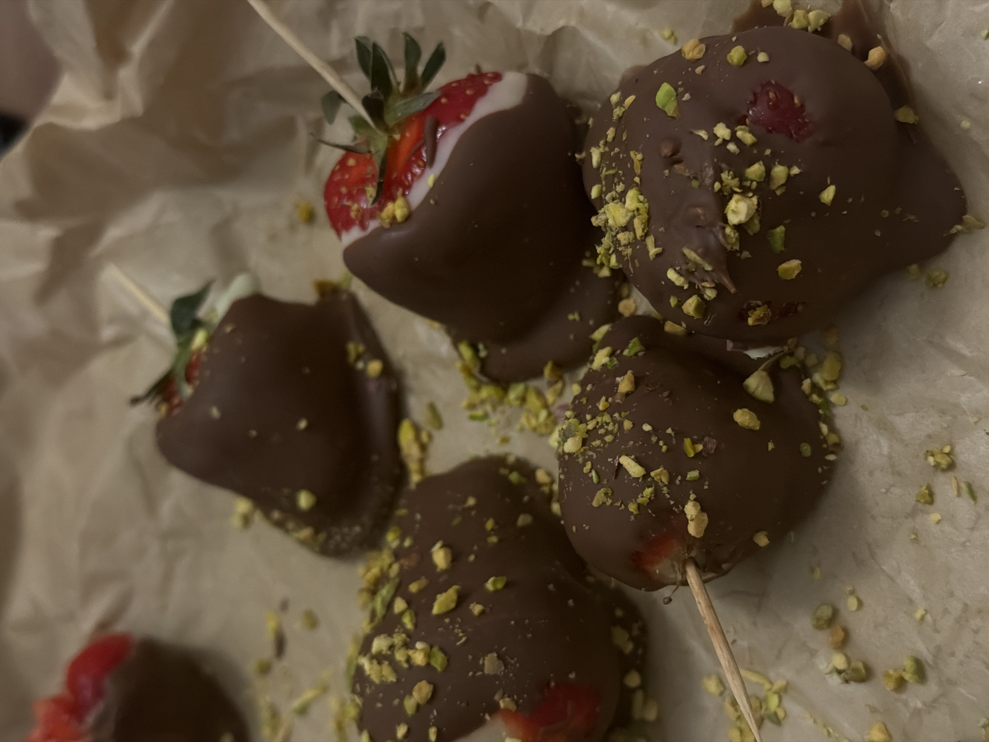 The image shows several chocolate-covered strawberries arranged on a sheet of parchment paper. Some strawberries are on sticks. One strawberry has a partial white coating under the chocolate. Crushed pistachios are scattered on several of the strawberries. It is unlikely that the image can be placed in Chaikovsky, Russia, based on the available information.