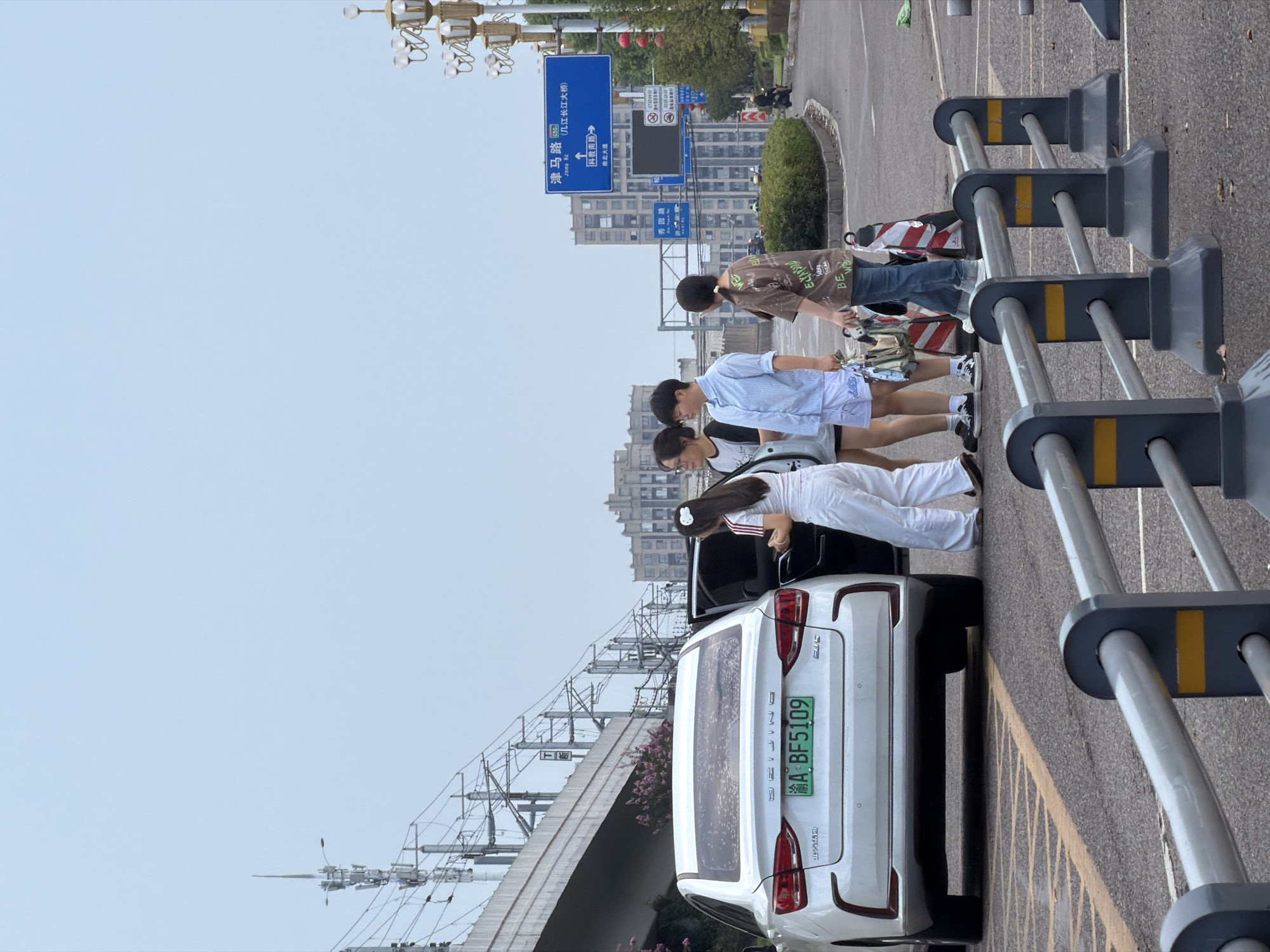 In Jiangjin, China, on a cloudy day, four young individuals stand near a white sedan. Two of the individuals are facing a black car, with one person opening the rear door. A third individual is looking at their phone, while the fourth person is looking away from the camera. The sedan's license plate reads "渝A BF5109". In the background, a train track overpass and buildings are visible. A blue road sign indicates "津马路" (Jinma Road) and "长江大桥" (Changjiang Bridge). A traffic sign with red lights is also present, along with a motorcycle rider in the distance. Safety cones are placed on the road, and a metal barrier is in the foreground.