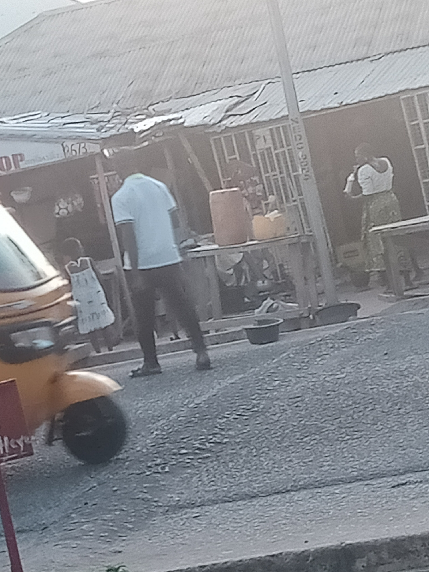 In Jalingo, Nigeria, a street scene unfolds with a yellow auto-rickshaw in the foreground on the left. The vehicle is parked on a paved road, partially obscured by the image's edge. Behind the auto-rickshaw, a young person in a light-colored dress stands near a stall. Further back, a man in a light blue shirt and dark pants walks on the road. A woman in a white top and patterned skirt is visible further in the background, near another stall. The stalls appear to be set up along the roadside, with corrugated metal roofs. A utility pole with writing on it stands between the man and the woman. The environment suggests a market or commercial area, with people engaged in daily activities.