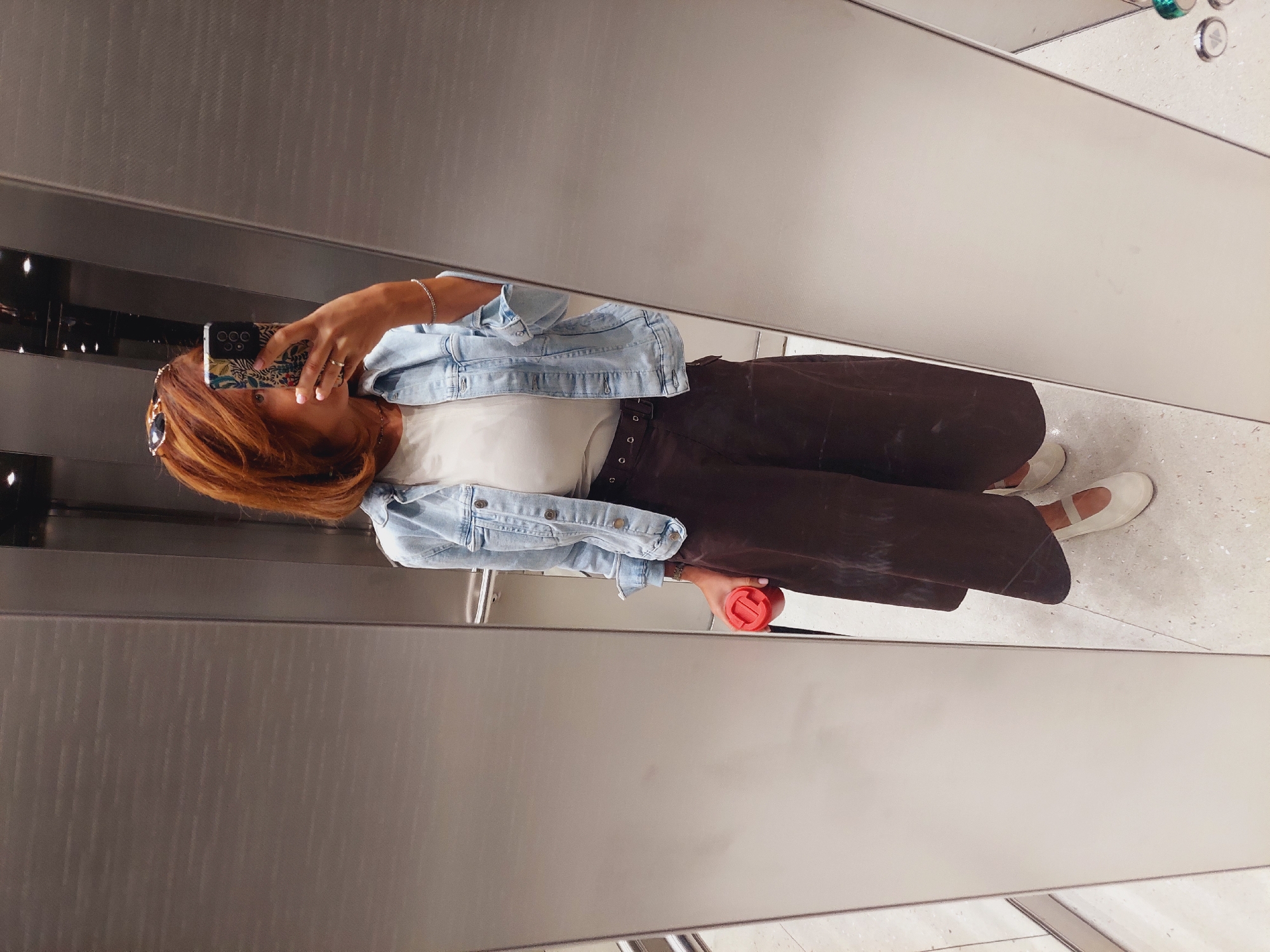 A woman with auburn hair, wearing sunglasses on her head, a light blue denim jacket, a white top, and dark brown wide-leg trousers, is taking a mirror selfie in what appears to be an elevator. She is holding a red travel mug in her left hand and a mobile phone in her right hand, which she is using to take the picture. The elevator has stainless steel walls, and the floor is tiled with a light, speckled pattern. Based on the metadata provided, the image was taken in Sofia, Bulgaria.