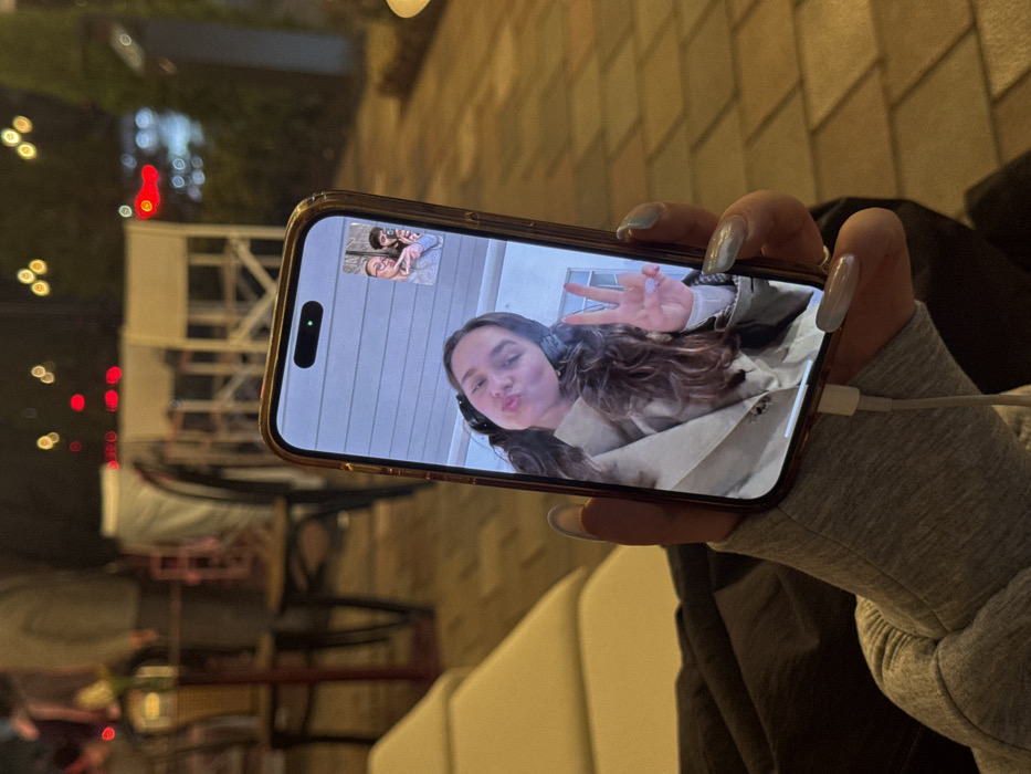 A right hand holds a vertically oriented smartphone, an iPhone model identifiable by its Dynamic Island feature, at night. The hand is clad in a grey, marled long-sleeved garment and displays long, manicured nails with a light-colored polish. A white charging cable is connected to the phone's port. The phone screen displays a video call. The primary video feed shows a young woman with dark, wavy hair, wearing headphones and a light-colored trench coat, making a "duck face" with pursed lips and flashing a peace sign with her left hand, which has light-colored nail polish. In the upper-right corner of the screen, a smaller video feed shows two additional individuals: one wearing glasses and making a peace sign, and another partially visible with dark hair. The background, largely out of focus, depicts an outdoor cafe or restaurant setting in the city of Yerevan, Armenia. Visible elements include blurred wooden chairs, some with white frames, dark tables, and indistinct figures of patrons. The ground is paved with light-colored square or rectangular tiles. Numerous blurred points of light, including prominent red and yellow hues, indicate ambient and decorative lighting, suggesting a nighttime scene.