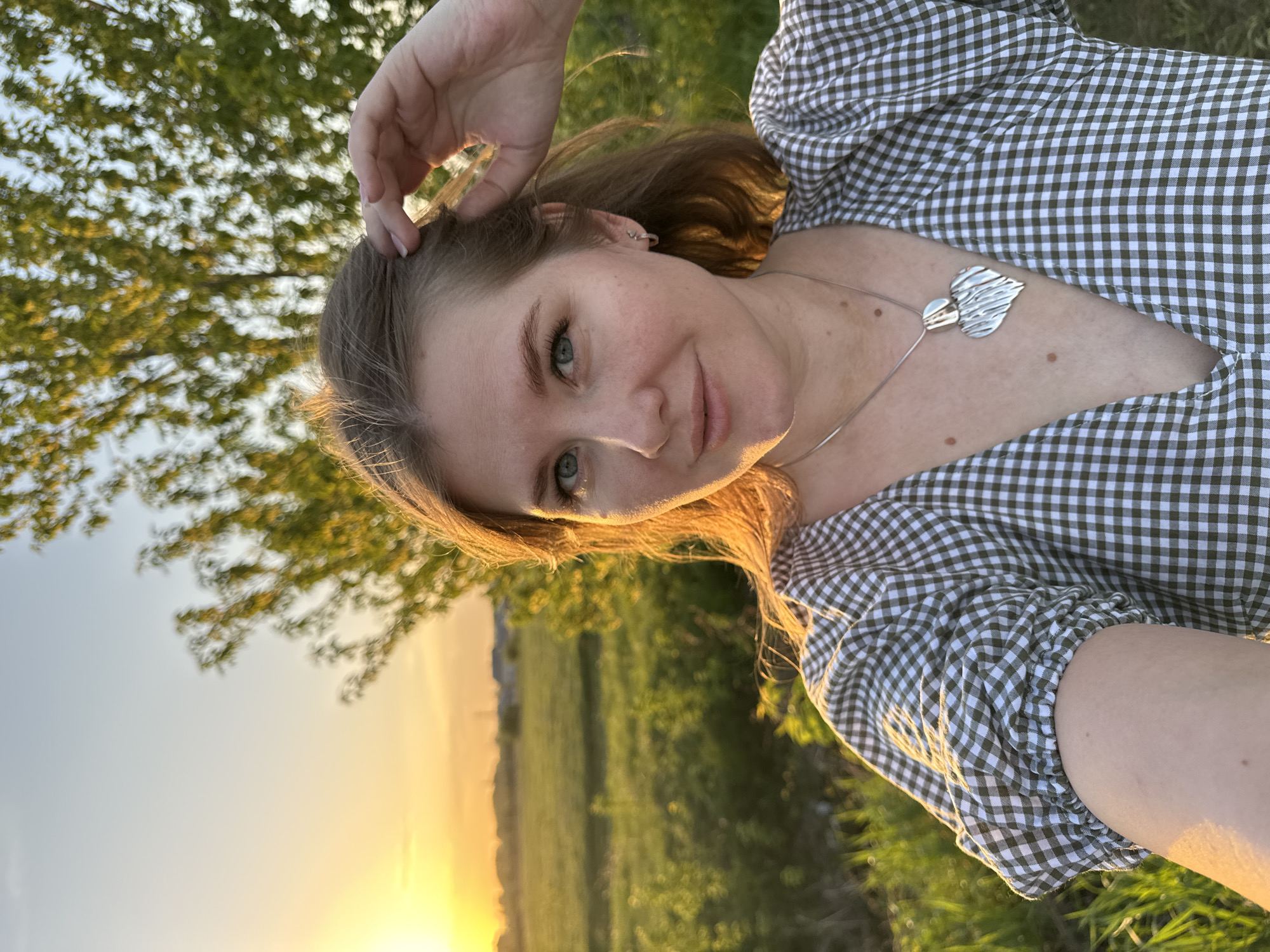 A woman is taking a selfie outdoors in the city of Горный, Russia. She is wearing a green and white checkered dress and a silver necklace with a heart-shaped pendant. Her hand is touching her hair. The background shows a field and trees, with the setting sun casting a warm glow.