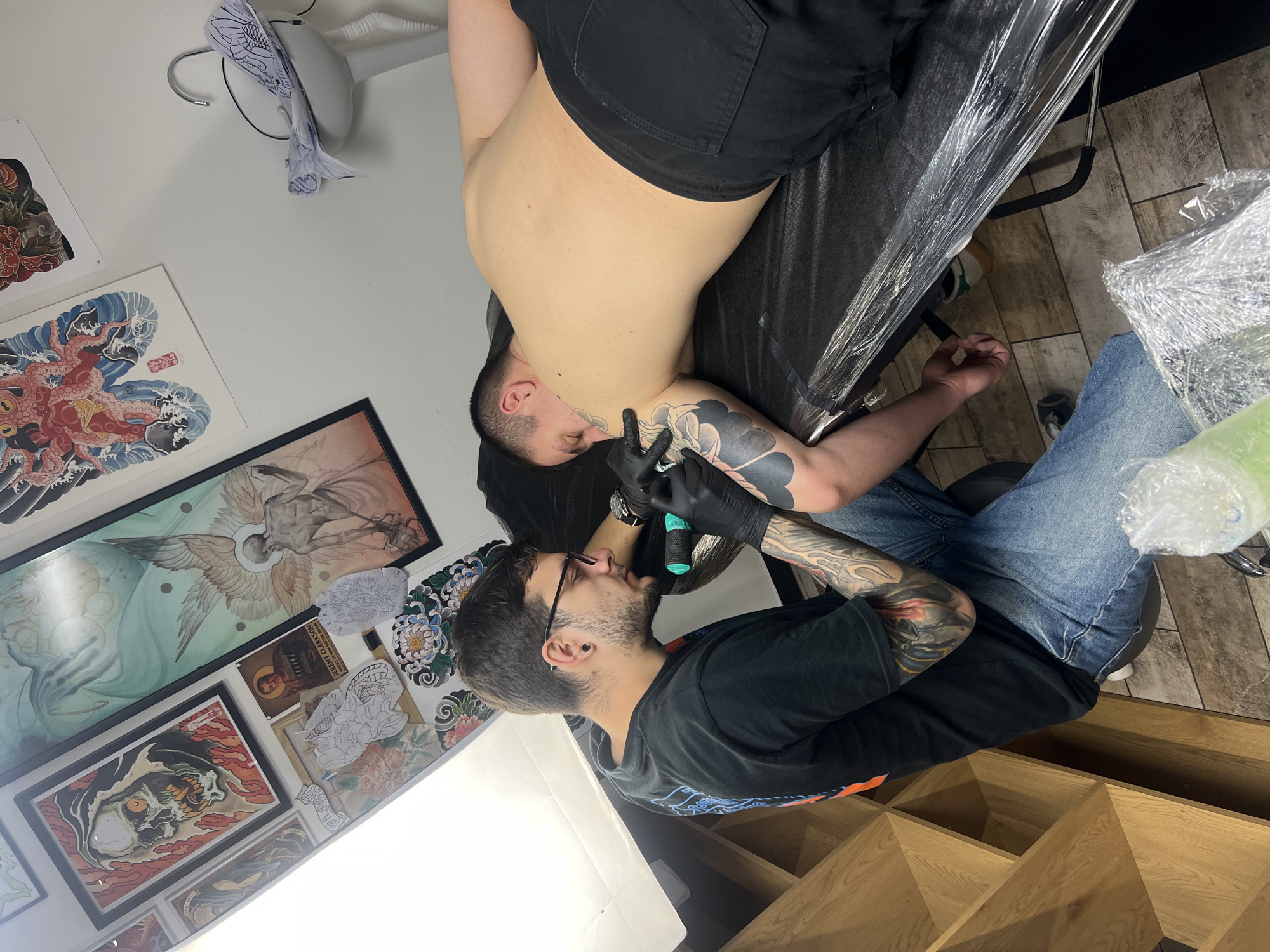 A tattoo artist is applying a tattoo on a customer's shoulder in what appears to be a tattoo parlor. The customer is lying on a covered tattoo bed. The tattoo artist, wearing glasses and a black t-shirt, is using a tattoo machine with black gloves. The artist's arms are covered in tattoos. The walls are decorated with tattoo art and sketches. There is bright light in the room, possibly from a lamp. The location is identified as the city of Sofia, Bulgaria.