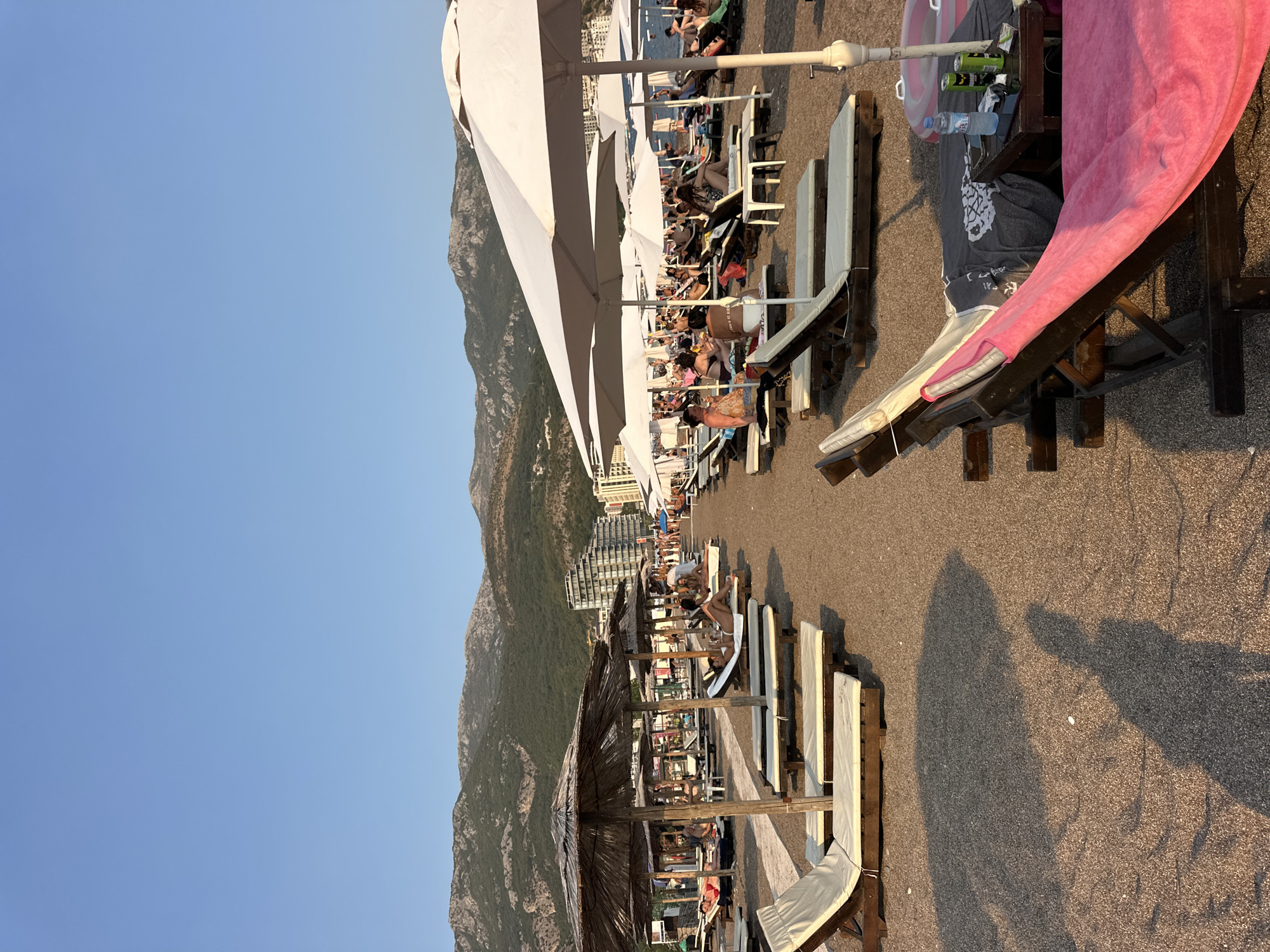This is a vertical, eye-level photograph of a crowded beach with mountains in the background. The scene depicts a sunny day, likely late afternoon or early evening, given the long shadows cast across the pebbly shore. The sky is a clear, bright blue, and the weather appears warm and pleasant.
The beach is lined with rows of white lounge chairs and umbrellas, some of which are large, white, fabric canopies, and others are made of straw. People are scattered throughout the scene, resting on the lounge chairs, some dressed in swimwear, while others appear to be walking or milling about. The activity is leisurely, with most individuals appearing relaxed and enjoying the beach atmosphere.
In the background, a large, modern hotel or resort complex is visible nestled against the base of steep, green-covered mountains. The mountains dominate the left side of the frame, extending upwards towards the sky. The sea is visible in the distance beyond the beachgoers.
In the foreground, on the right side of the image, a dark wooden structure with a pink towel draped over it is partially visible. Scattered on the structure are a few bottles and cans. Shadows of people are prominent on the beach, suggesting the sun is low in the sky. The overall mood of the image is one of a typical summer day at a popular tourist destination. No visible text is present in the image.