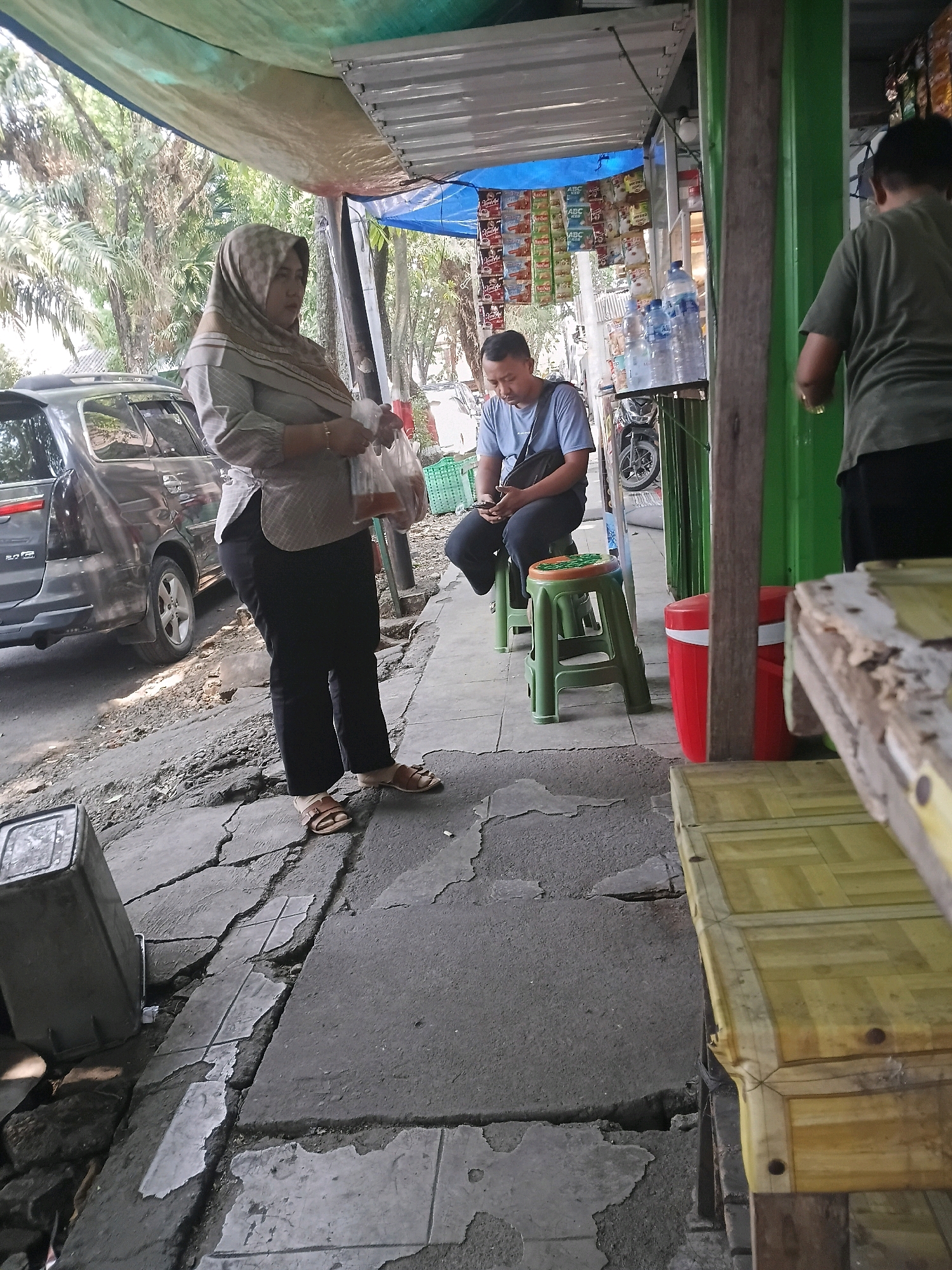 The image depicts an outdoor street scene in Purwodadi, Indonesia, likely in front of a small shop or food stall. In the foreground, a woman wearing a patterned hijab and a light-colored blouse paired with black pants stands on a cracked concrete sidewalk. She is holding a clear plastic bag containing food or drinks. To her right, a man in a blue t-shirt and black pants sits on a green plastic stool, looking down at his mobile phone. Behind him, a display of packaged goods hangs from a structure. Further to the right, another person wearing a green t-shirt and black pants stands facing away from the camera. A dark gray SUV is parked on the left side of the scene, with part of its rear visible. The environment includes a roofed structure with what appears to be a blue tarp and corrugated metal, overhead foliage, and various plastic containers. The overall impression is a casual, everyday scene on a street in a warm climate.