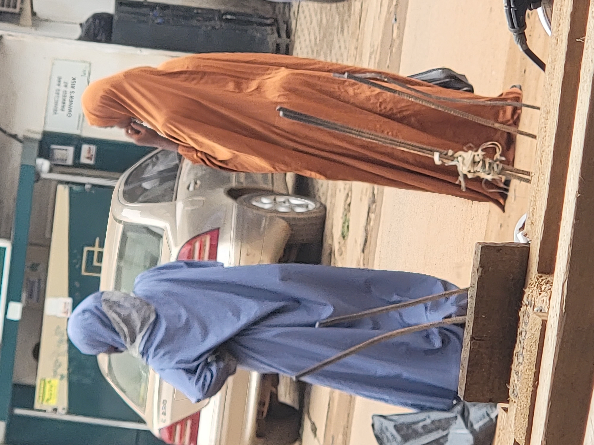 Two people, each wearing a full-body covering, are carrying long, slender objects. One person is wearing an orange covering, and the other is wearing a blue covering. They are near parked cars in an urban environment. Based on the prompt, the location is Maiburiji, Nigeria. The photograph is taken outdoors during daylight. The exact action or purpose of carrying the slender objects is not evident.