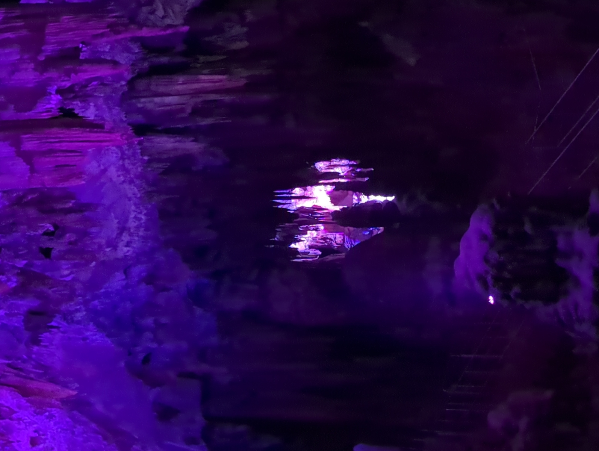 The image depicts the interior of Ledenika Cave, a karst cave located near the city of Vratsa, Bulgaria. The cave is illuminated with vibrant purple lights, highlighting the dramatic stalactites and stalagmites that adorn the cave's ceiling and floor. In the foreground, a path with a railing guides visitors through the cavern. The lighting creates an ethereal atmosphere, casting deep shadows and emphasizing the natural formations. The scene is devoid of people, suggesting a quiet moment within the cave. The overall impression is one of natural beauty amplified by artificial illumination, showcasing the geological wonders of Ledenika Cave.