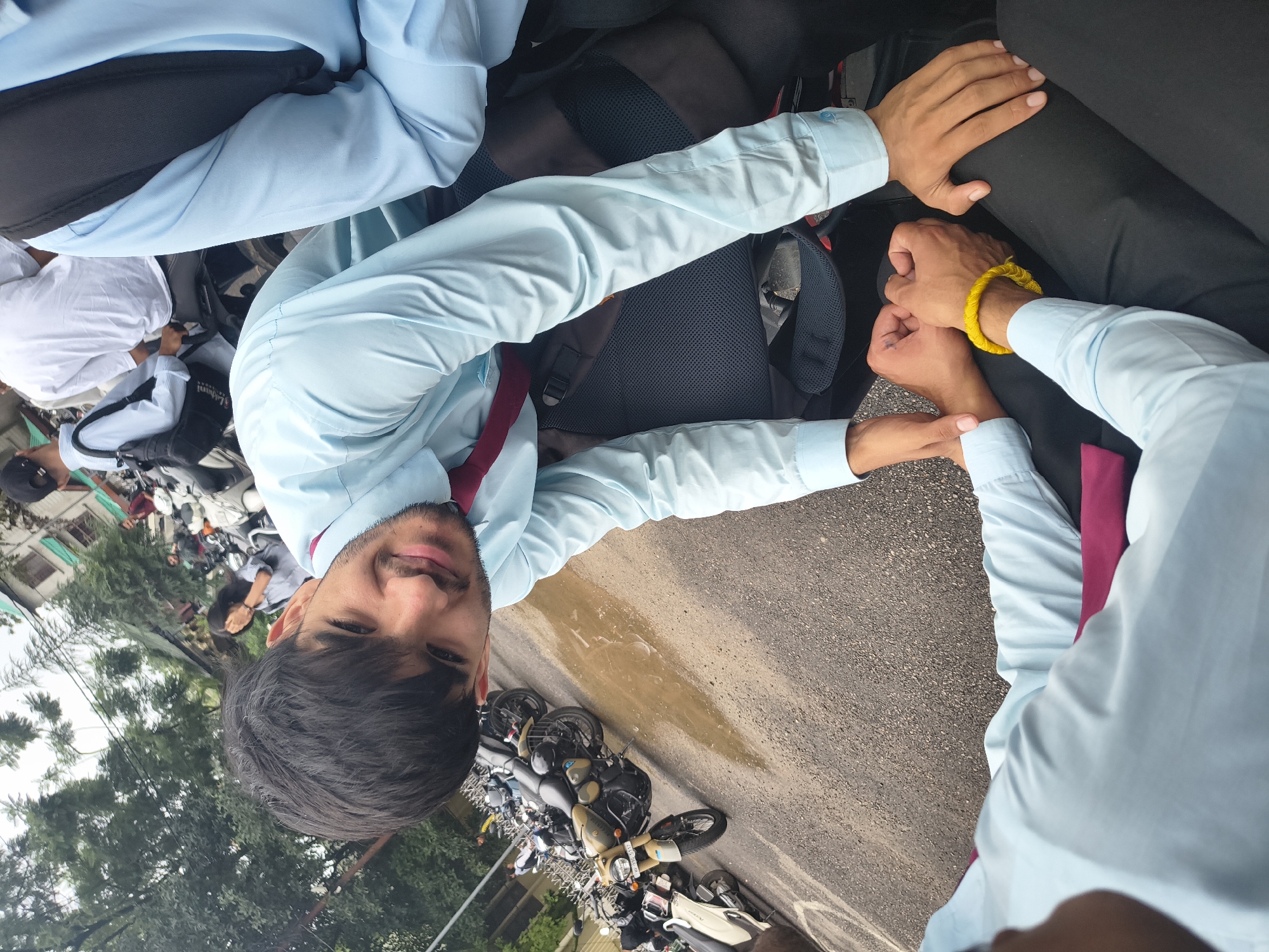The image depicts a street scene in Dharamshala, India, on a cloudy day. Several young men, presumably students, are gathered around motorcycles. The foreground is dominated by two individuals wearing light blue shirts and dark pants, with one of them sporting a maroon tie and a yellow bracelet. Their hands are clasped, suggesting an interaction or perhaps a playful gesture. In the background, a line of motorcycles is parked on a wet asphalt road, with puddles indicating recent rain. One motorcycle, a tan and black Royal Enfield, is prominently featured. Beyond the motorcycles, trees and buildings are visible, hinting at an urban or semi-urban environment. The overall atmosphere appears casual, possibly a break or gathering before or after an event.