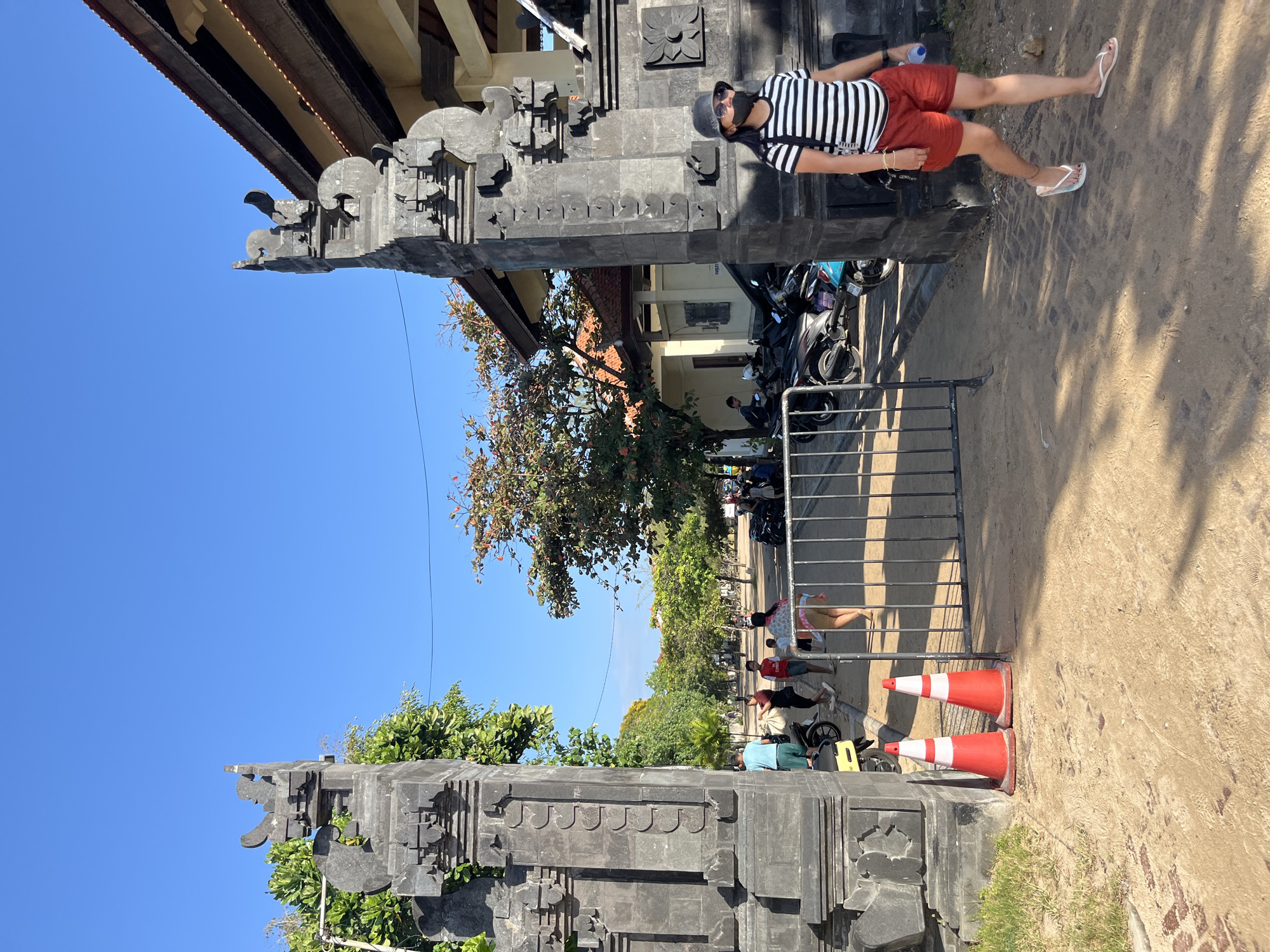 In Kuta Selatan, Indonesia, the image captures an outdoor scene under a clear blue sky. The foreground features two red and white traffic cones placed on a sandy path, with a metal barrier behind them. To the left, a large stone structure, part of a traditional Balinese gate, stands prominently. On the right, a woman wearing a black and white striped shirt, red shorts, a black hat, sunglasses, and a black face mask walks away from the viewer, carrying a dark bag over her shoulder. She is wearing flip-flops. Behind the traffic cones and barrier, several people are visible, some walking and others near parked motorcycles. Trees line the path, and a building with an orange tiled roof is visible in the background. The overall impression is a sunny day at a public entrance or pathway, likely in a tourist area given the architecture and attire.