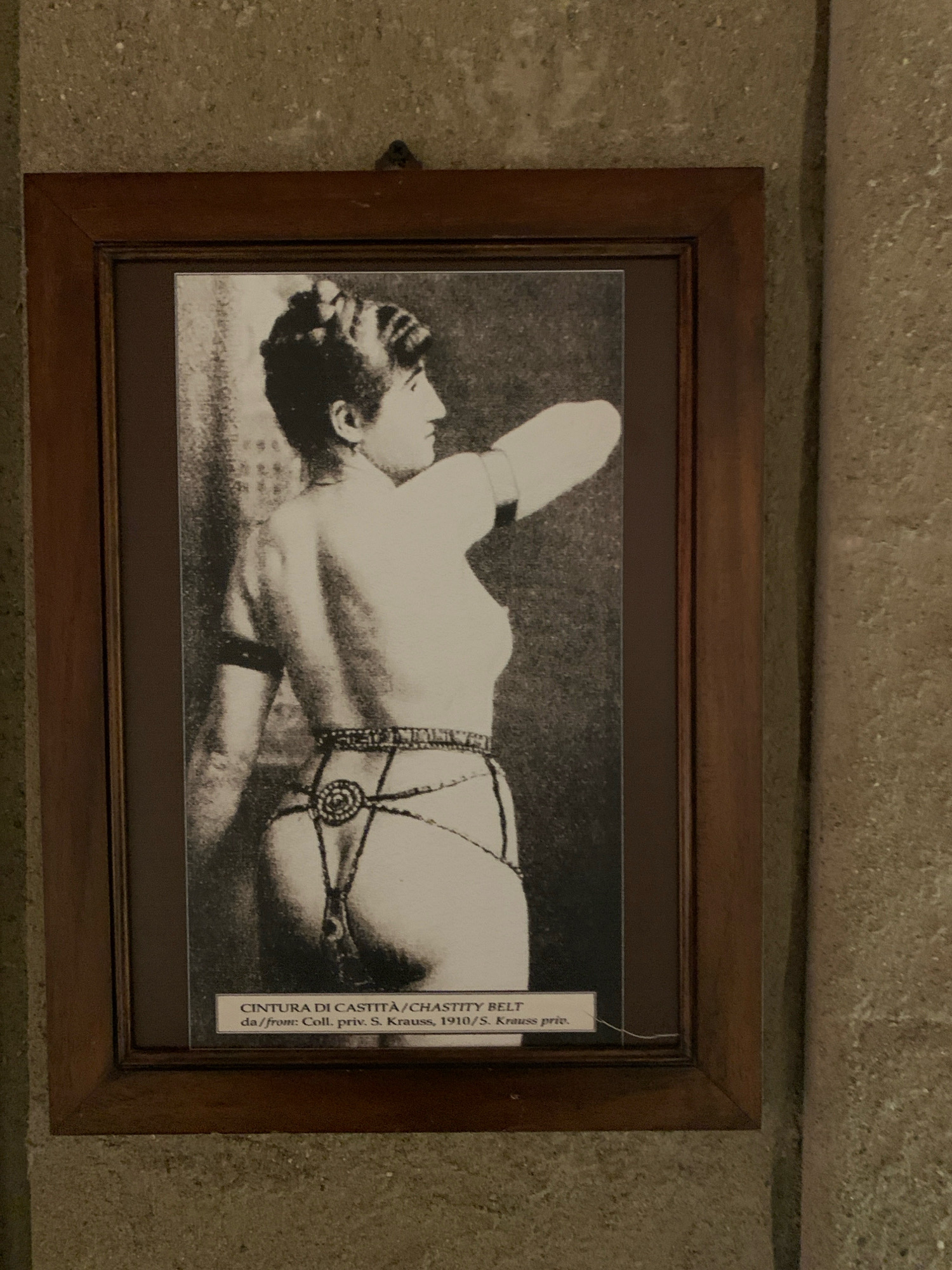 A framed black and white photograph depicts a woman wearing a chastity belt. The photograph is hanging on a textured stone wall, with a portion of a pillar visible on the right. The woman is shown in profile, facing right, with her arms raised. She has her hair styled up. The chastity belt appears to be made of metal and fabric. Below the photograph, a label reads "CINTURA DI CASTITÀ/CHASTITY BELT" and "da/from: Coll. priv. S. Krauss, 1910/S. Krauss priv.". The photograph is housed in a wooden frame. This image is located in the Torture Museum in the City of San Marino, San Marino.