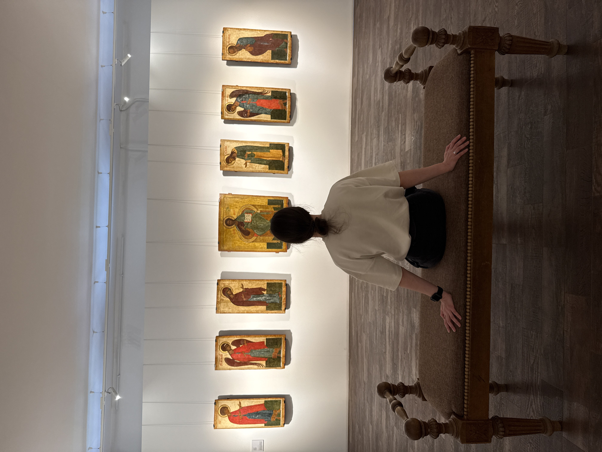 A person sits on a bench in an art gallery in Nizhny Novgorod, Russia, viewing seven religious icons hanging in a vertical arrangement on a white wall. The person has dark hair pulled back, wears a light-colored top, and dark pants. The bench is ornate, with carved wooden legs and a fabric seat. The floor is made of wood planks. The icons are illuminated by track lighting near the ceiling.