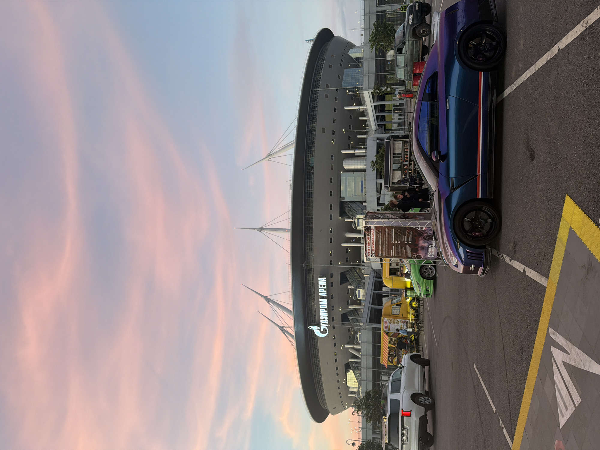 The image shows the Gazprom Arena in St. Petersburg, Russia, during a sunset. The sky is filled with soft pink and blue hues, indicating the time of day. In the foreground, a dark blue Nissan GT-R with a multi-colored stripe on its side is parked in a lot. Other vehicles, including a white SUV and a green sports car, are also present, as well as a yellow food truck. Several people are visible, some standing near the food truck and others near the entrance of the stadium. The stadium itself is a large, modern structure with a distinctive roof design. The overall scene depicts a lively atmosphere outside a major sporting venue.