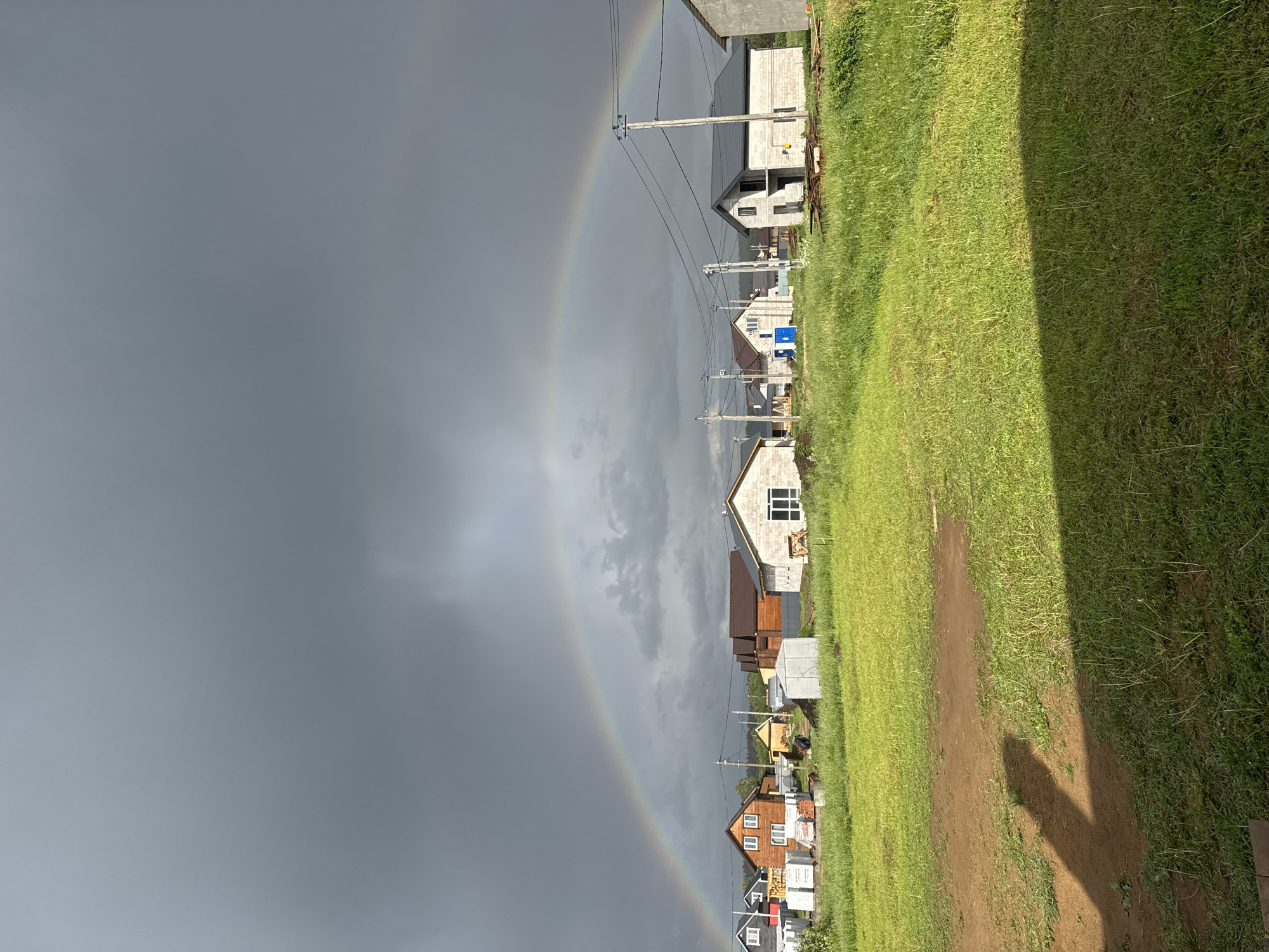 The image depicts a residential area under a dramatic sky exhibiting a full rainbow. The location is Ферма, Russia, as indicated by the surrounding environment and typical architectural styles.
Several residential houses are lined along a green expanse that appears to be a wide pathway or field. These houses are of various construction stages, from complete to under construction, indicating ongoing development or varying levels of completion. Power lines and poles are visible, running parallel to the houses.
The sky occupies a significant portion of the photograph and shows a stark contrast between a dark, overcast expanse and a bright, complete rainbow. The rainbow's arc seems to originate and terminate within the depicted area, enhancing the visual impact of the scene.
The pathway or field consists of green grass and patches of bare earth, suggesting regular use or uneven growth. The lighting is bright, although the overcast sky suggests it is not direct sunlight. The houses are simple structures with various roofing materials and designs. The overall impression is of a typical suburban residential landscape in Russia experiencing dramatic weather conditions.