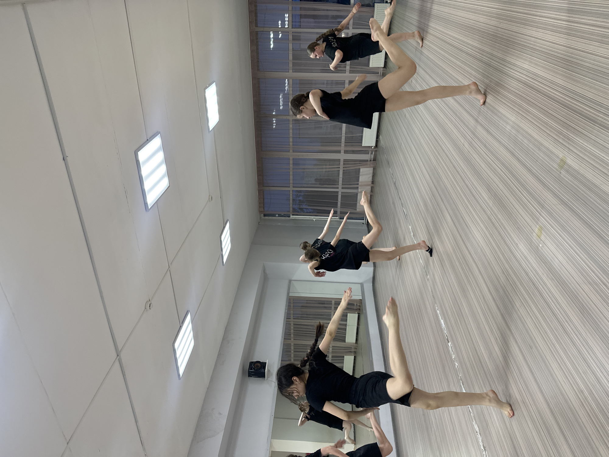 The image depicts a dance studio in Votkinsk, Russia. A group of young female dancers, dressed in matching black shorts and t-shirts, are practicing a contemporary dance routine. They are positioned in a line, with each dancer mirroring the pose of the others. The studio has a light-colored wooden floor and white walls with rectangular ceiling lights. There is a window with horizontal blinds in the background.