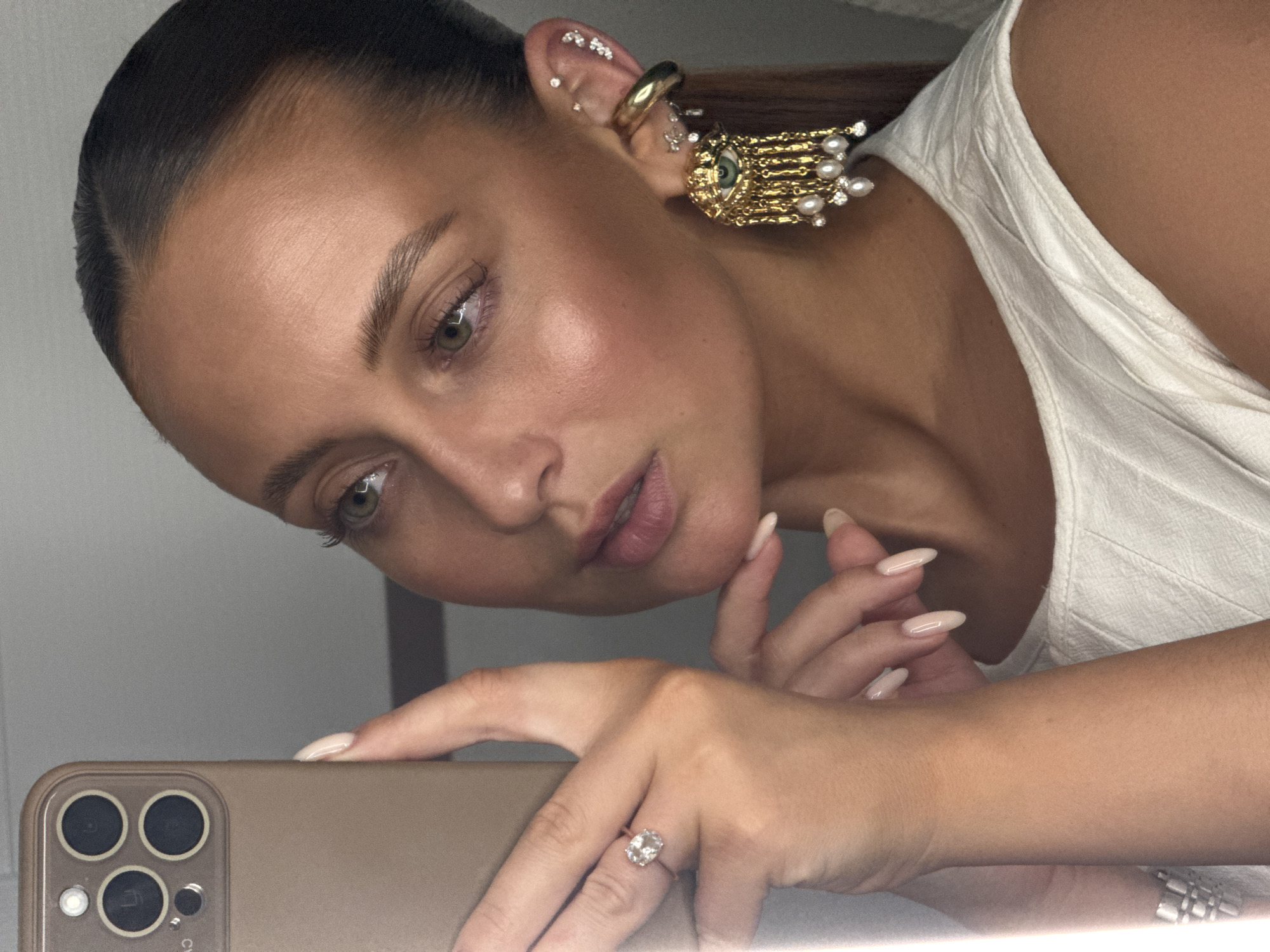 A woman is taking a selfie with a tan iPhone, focusing on her face, ear jewelry, and hands. She has slicked-back hair, light skin, and wears an elaborate gold earring with a small eye and dangling pearls on her left ear. Multiple smaller piercings are also visible. She wears a large diamond ring on her left ring finger. She wears a white top. The location in the city of Chaikovsky, Russia, is not apparent from the image.