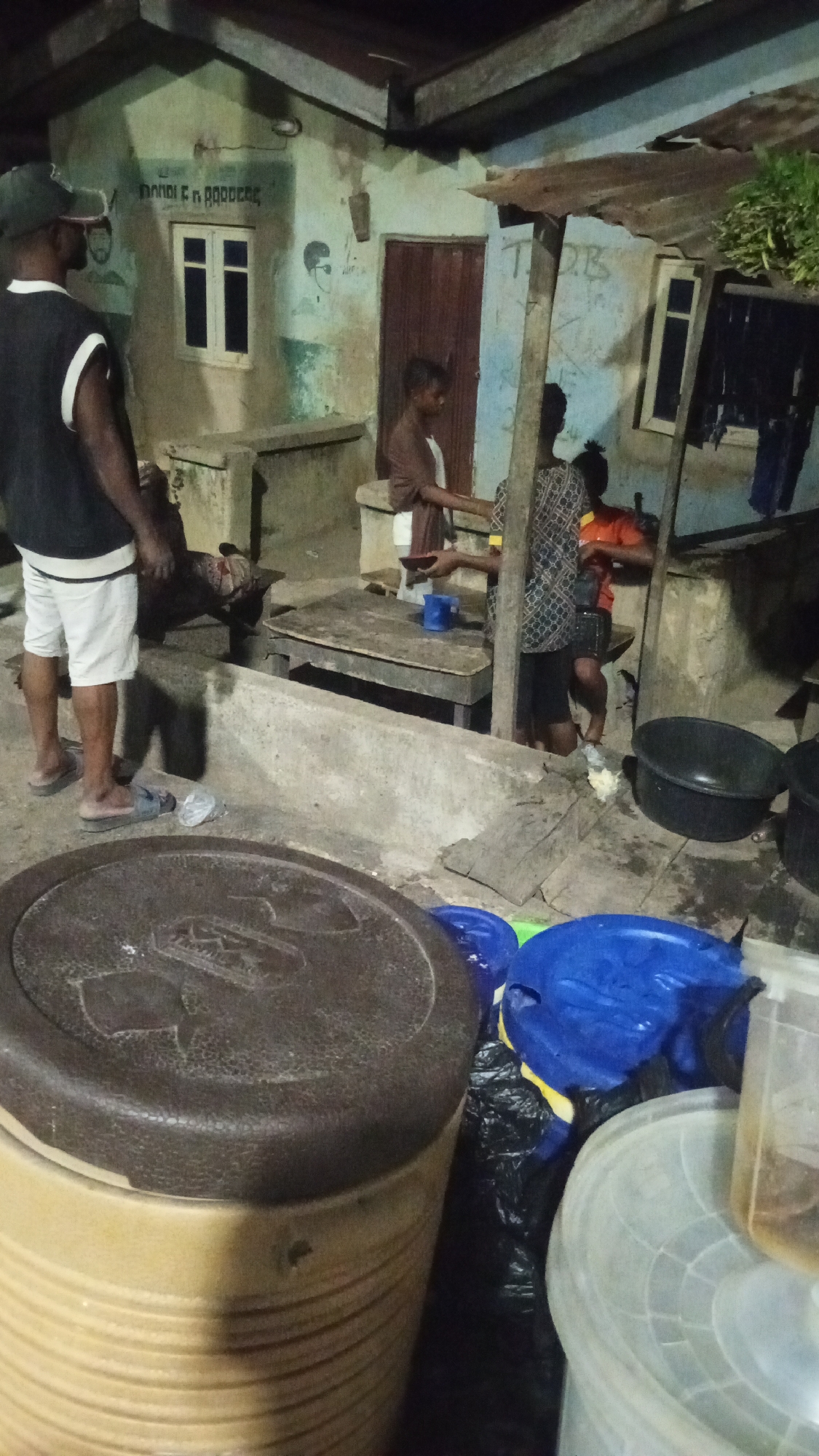 In Adankolo, Nigeria, a group of people are gathered outside a weathered building at night. A man wearing a black vest and white shorts stands on the left side of the image, while three other individuals are near a wooden table. One is handing a blue cup to another, while the third sits nearby. Several black basins and a large, brown-lidded container are in the foreground. The building has faded writing and drawings on the walls, and a window and a door are visible. The lighting is dim, creating a muted and somewhat grainy visual.