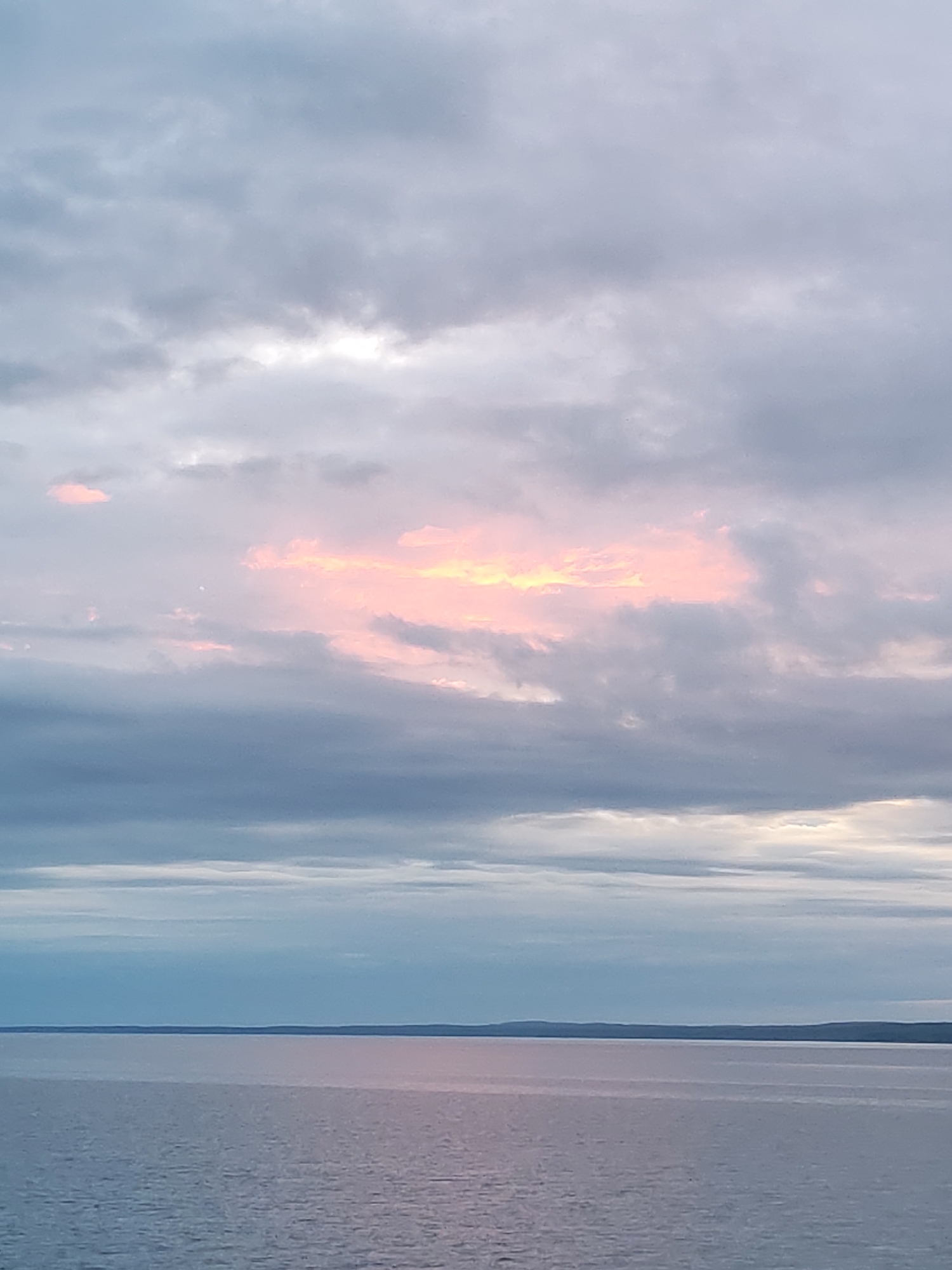 The image captures a serene landscape featuring a vast body of water under a dramatic sky, likely at sunrise or sunset. The upper two-thirds of the frame are dominated by a cloudy sky. Overcast clouds in shades of muted blue, gray, and lavender stretch across the expanse. A prominent horizontal band of soft, luminous pink and orange light breaks through the clouds in the mid-upper section, suggesting the sun's presence below the horizon or obscured by clouds. This vibrant band contrasts with the cooler tones of the surrounding cloud cover.
The bottom third of the image shows a calm body of water, possibly a large lake, sea, or ocean. The water surface reflects the muted colors of the sky, appearing a soft blue-gray with hints of pink where the light from the clouds touches it. Gentle ripples are visible across the water, indicating a tranquil environment. On the distant horizon, a low, dark landmass stretches across the entire width of the frame, providing a clear boundary between the water and the distant background. There are no visible people, animals, man-made structures, or text in the scene. The overall impression is one of natural beauty and tranquility during a transitional time of day.