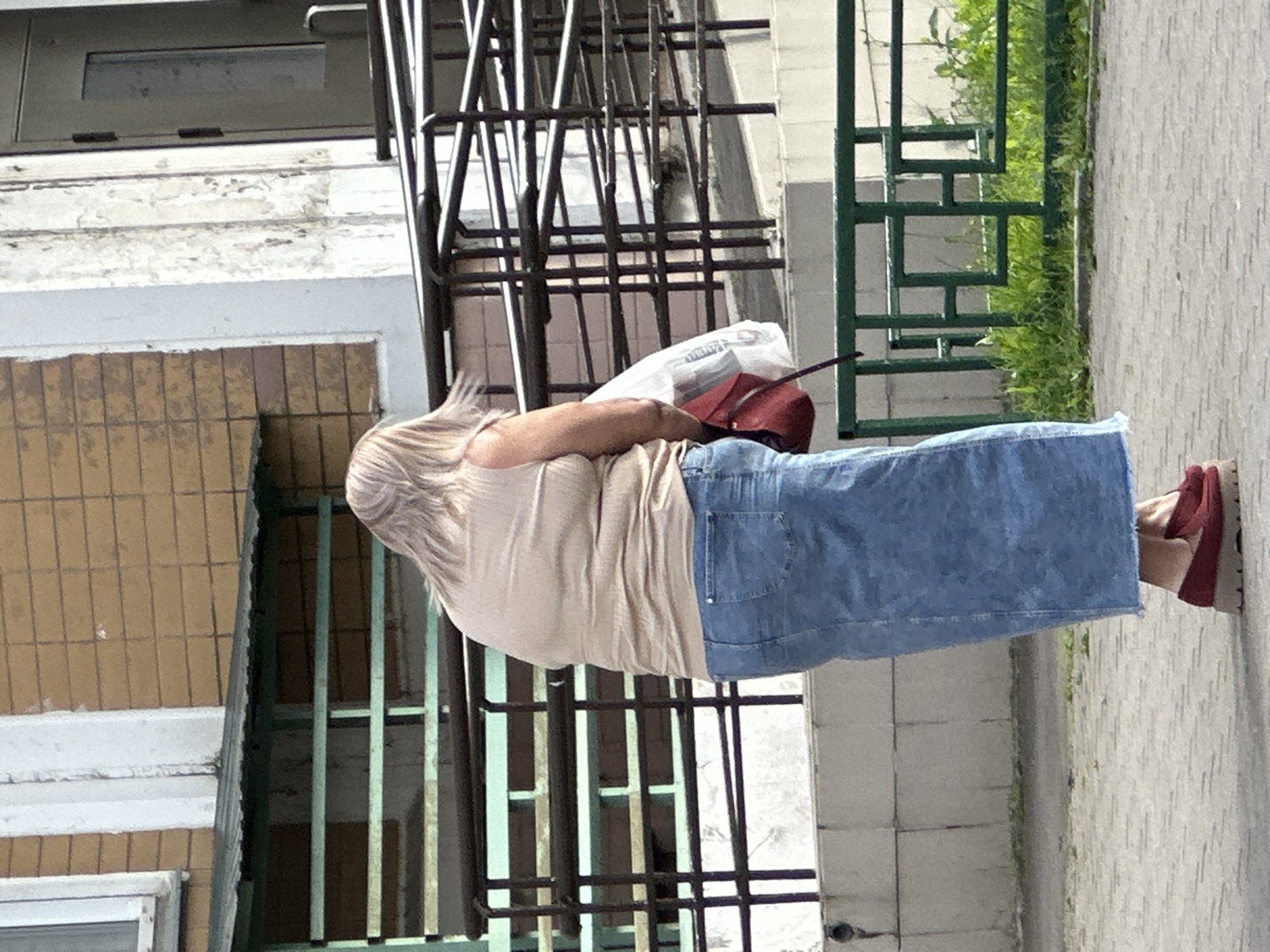 A woman with blonde, windswept hair is standing in front of a brick building with a white column. She is wearing a beige sleeveless top, a long denim skirt, and dark red platform loafers. She is holding a red purse and a white plastic bag in her left hand. The background includes a green metal railing and lush green grass. The scene appears to be outdoors, possibly in the city of Podolsk, Russia, given the architectural style of the building.