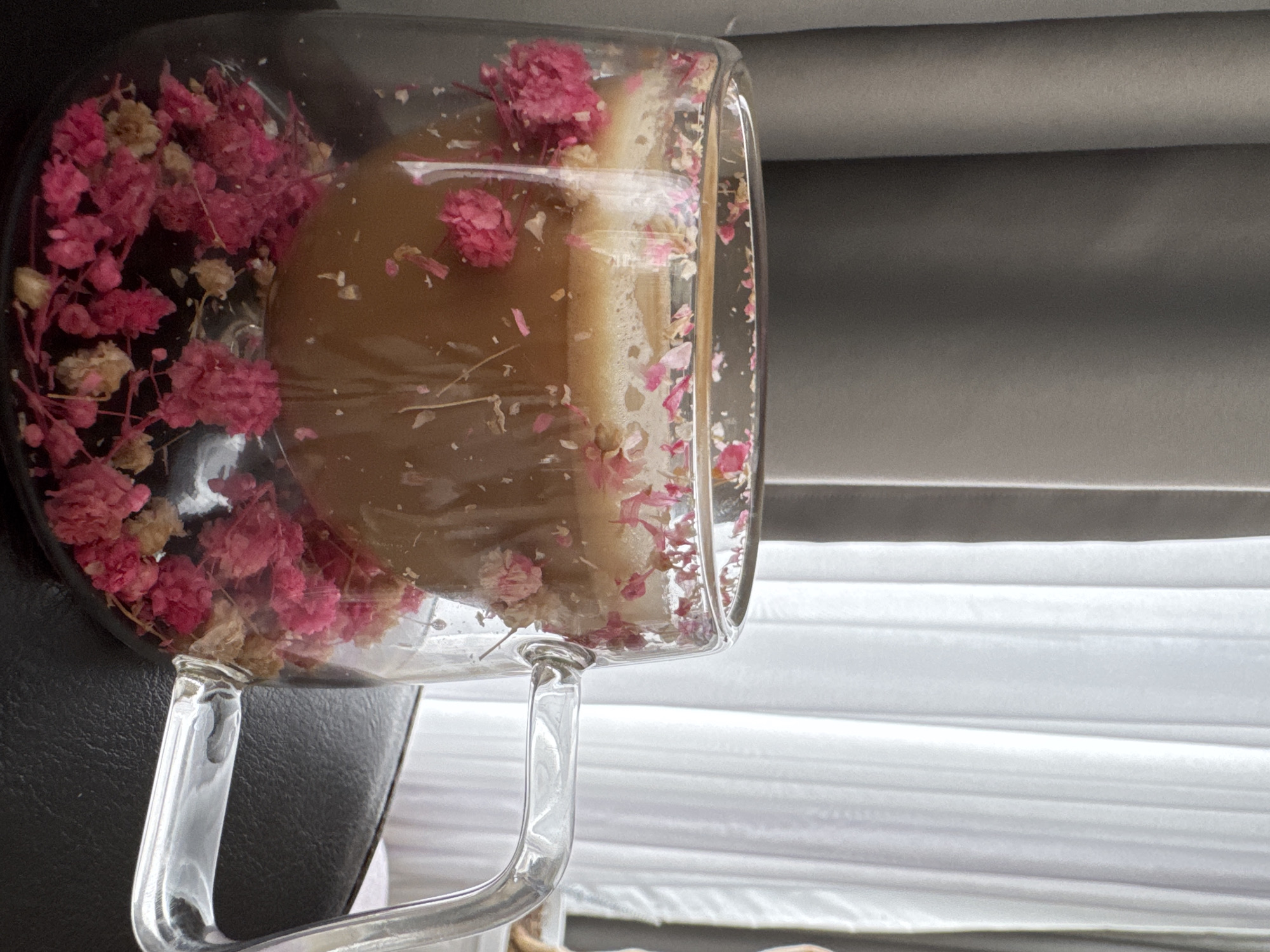 A glass mug filled with a tan liquid and pink flowers sits in front of a window with white blinds and gray curtains. The mug is on a black surface. The location is Varna, Bulgaria.