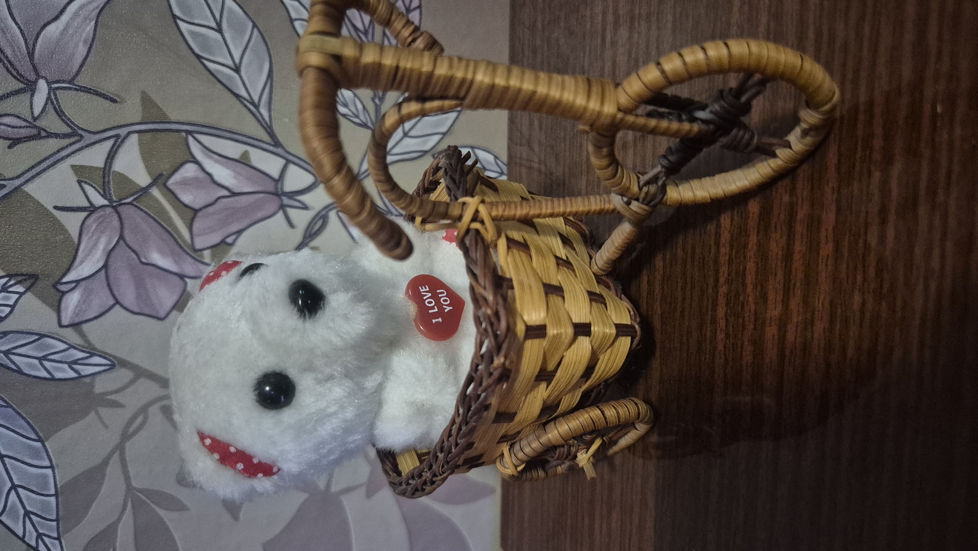 A white plush teddy bear with black button eyes and red polka dot accents on its ears is seated in a woven basket attached to a miniature woven bicycle. A small, heart-shaped red sticker with the words "I LOVE YOU" is affixed to the bear's chest. The bicycle and basket are made of woven natural fibers, likely wicker or rattan, and have a rustic appearance. The background features a floral wallpaper with muted purple and gray tones. The scene is set on a dark wooden surface. There are no people present. The location of Butovo, Bulgaria, is not evident in the image itself.