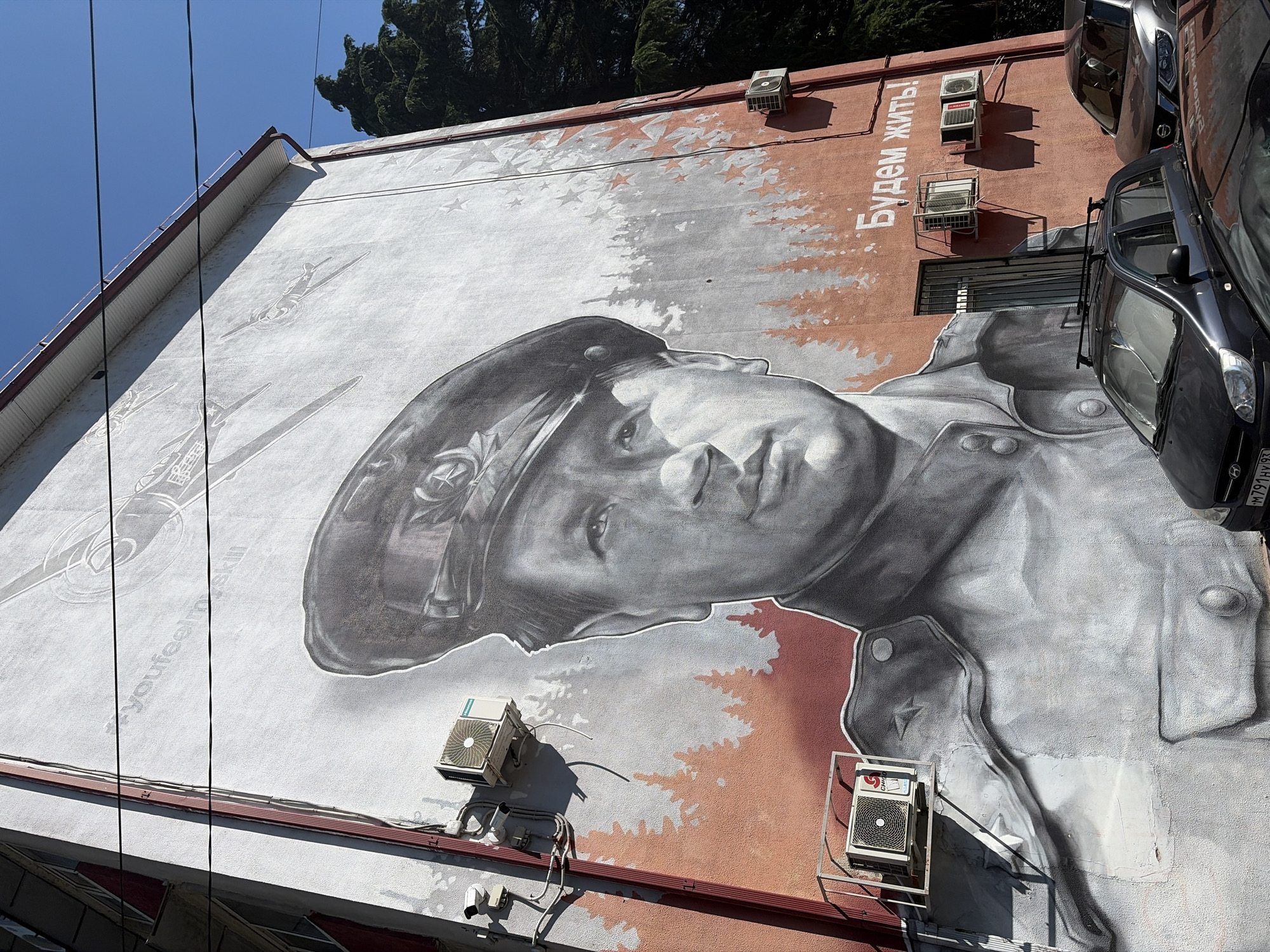 This is a photograph of a large mural painted on the side of a building in Sochi, Russia. The mural depicts a black and white portrait of a man in a military uniform and cap, looking slightly upwards. He appears to be a historical figure, possibly a soldier or leader. The mural is rendered in grayscale with some hints of reddish-brown paint in the background, resembling a stylized forest or camouflage pattern. Scattered stars are also visible in the background.
The mural is on a tall building with windows and several air conditioning units mounted on the exterior wall. The building has a reddish-brown facade in some areas, contrasting with the white and gray of the mural. There are power lines running in front of the mural.
In the upper right of the image, parts of two cars are visible parked near the building, one dark gray and another lighter, possibly silver. The sky is a clear, bright blue, suggesting a sunny day. The trees visible at the top of the image appear to be tall and dark green.
There is text visible on the building in white Cyrillic letters that reads "Будем жить!". This translates to "We will live!". There is also some fainter text in English on the mural itself, part of which reads "*youfeel"...skin".
The overall impression is one of remembrance and possibly a message of resilience or hope. The scale of the mural suggests it is a prominent public artwork. The time of day appears to be midday, given the bright sunlight and distinct shadows cast by the air conditioning units.