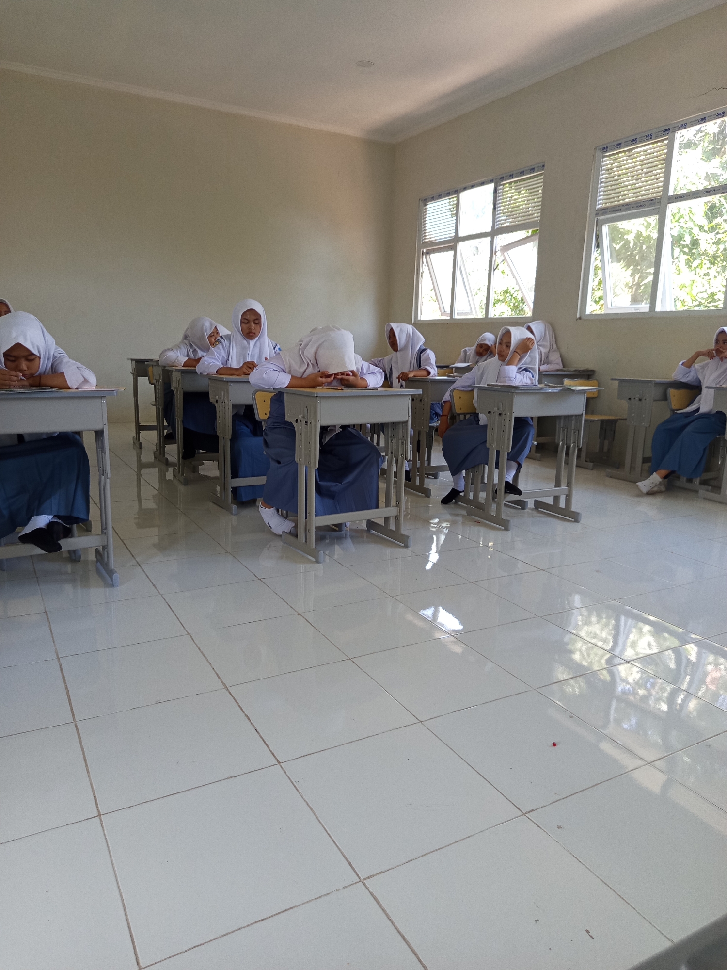 In a classroom in Lumutan, Indonesia, several female students in white hijabs and blue uniforms sit at their desks, some appearing to be asleep or resting their heads on their arms. The room features tiled flooring and large windows with blinds, allowing natural light to enter. The desks are simple, metal-framed tables with attached seats. The overall impression is one of a typical school day, possibly a quiet moment during a lesson or a break.