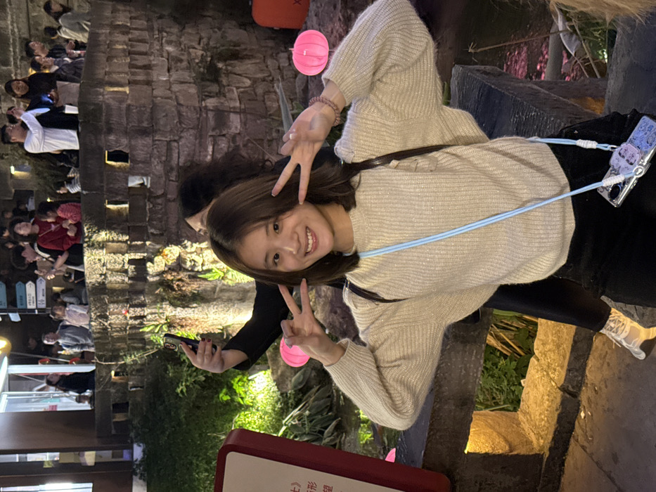 This image captures a nighttime outdoor scene in a public recreational area within Nan'an District, China, featuring multiple individuals and stone architectural elements under artificial illumination.
In the immediate foreground, a young East Asian female with shoulder-length dark brown hair stands facing the camera, smiling with teeth visible, and making a "peace sign" gesture with both hands. She wears a light beige knitted sweater with subtle shimmer and ruffled shoulder details, and dark pants. A light blue cord functions as a phone lanyard, holding a clear phone case containing a purple-patterned smartphone adorned with small charms. A dark strap from an unseen bag is visible over her right shoulder. Her right elbow rests on a dark stone railing or ledge.
Partially obscured behind and to the left of the primary subject, another individual is seen holding a dark-colored smartphone vertically, appearing to take a photograph or video.
The background consists of two distinct levels. At the lower level, behind the foreground railing, a dark body of water reflects artificial lights, bordered by illuminated green foliage. Two pink, spherical lanterns are visible on the right. A red rectangular sign with white Chinese characters is partially visible on the far left.
The upper level features an elevated stone pathway or bridge, bordered by a substantial stone wall with rectangular cutouts, which is illuminated by warm spotlights. On this pathway, approximately fifteen individuals, including adults and children, are present. Many are observing the scene, with several holding smartphones, indicative of photographic activity. A multi-directional signpost with Chinese characters is positioned on the far upper left, adjacent to a section of a building with lit windows.