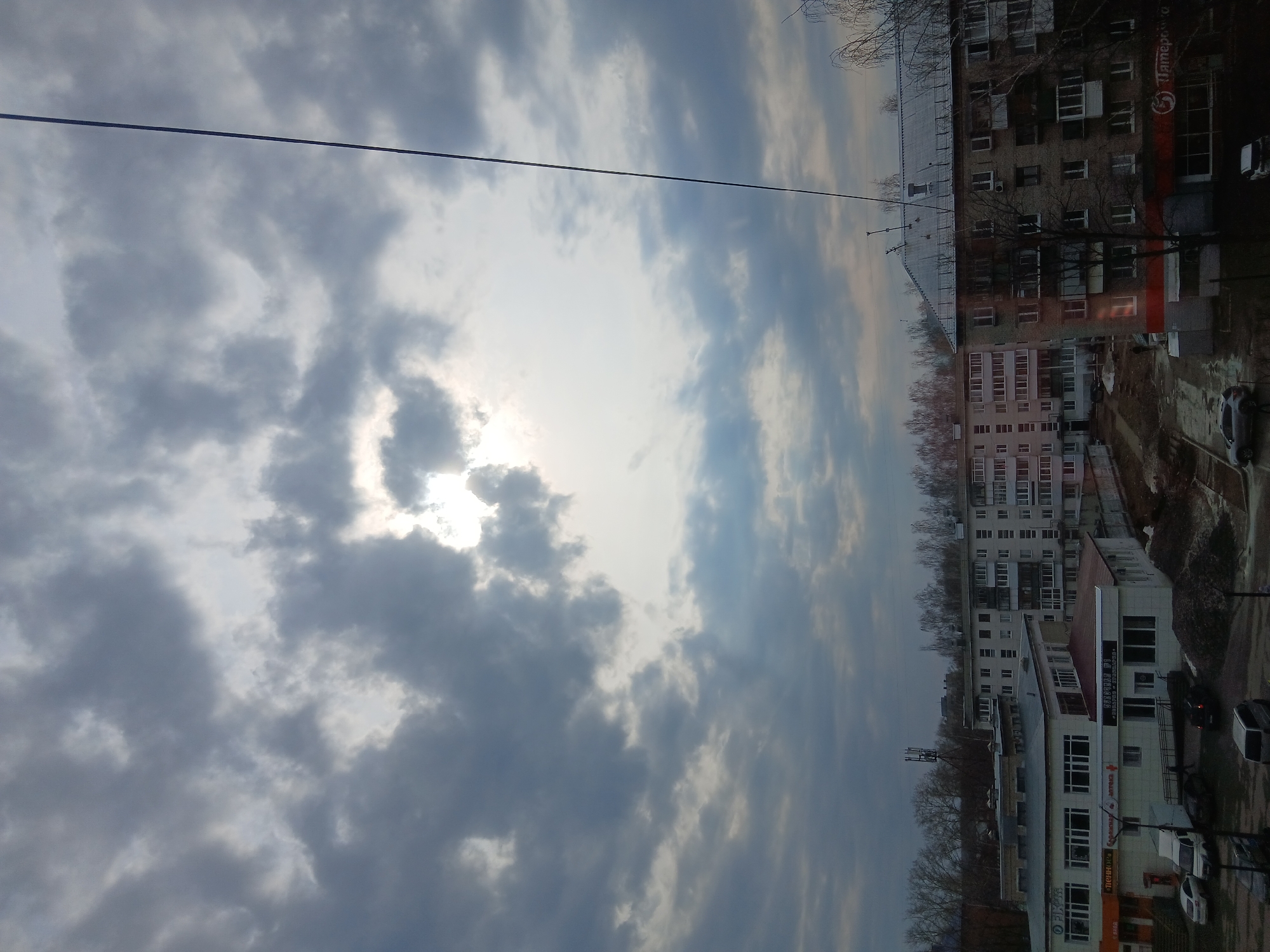 The image depicts a cityscape in Chaikovsky, Russia, under a cloudy sky with the sun obscured. The buildings are a mix of residential and commercial structures, varying in height and color. A prominent brick building has a sign in Russian. A power line stretches across the top portion of the image. Some vehicles are parked on the streets below. The environment suggests an overcast or winter day.