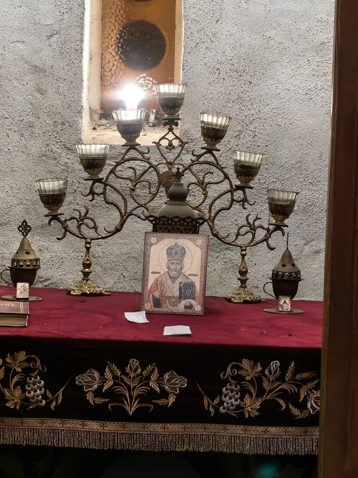A still, indoor shot presents an altar-like setting, featuring a red-covered table adorned with a complex candelabra, religious icon, and other articles. The candelabra, crafted from bronze, holds seven clear glass candle holders arranged symmetrically across its ornate, looping arms. In the center, a religious icon stands prominently, depicting a man with a halo and a book, dressed in traditional religious attire. Adjacent to the candelabra are two incense burners, each constructed from dark metal with delicate cross patterns, resting on metal saucers. A book lays to the left of the icon. The table is covered with a deep red cloth, edged with a black strip adorned with gold embroidery depicting floral and vine motifs. Beneath the cloth, a fringe of gold tassels adds texture. The scene is set against a rough, grey wall, with a small window above, its interior covered, reflecting light back into the room. A vertical wood structure partially frames the scene on the right. The location is Unknown, Unknown.