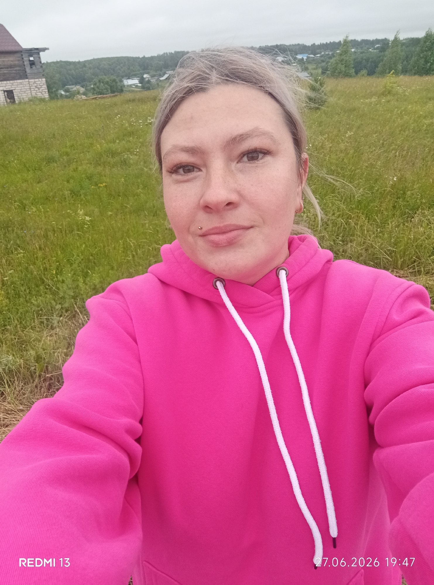 This is a selfie of a woman wearing a bright pink hooded sweatshirt. She has light-colored hair, some freckles on her face, and a small metal piercing on her lip. The background shows a grassy, slightly overgrown field with some trees and buildings visible in the distance. The sky is overcast, suggesting a cloudy or misty day. In the far left background, there is a partially constructed wooden and brick building. The bottom right corner of the image displays a timestamp: "27.06.2026 19:47". The bottom left corner displays "REDMI 13", indicating the device used to capture the photo. The setting appears to be rural, consistent with the provided location context of Фоки, Russia. The woman's expression is neutral, and the overall mood is relaxed.