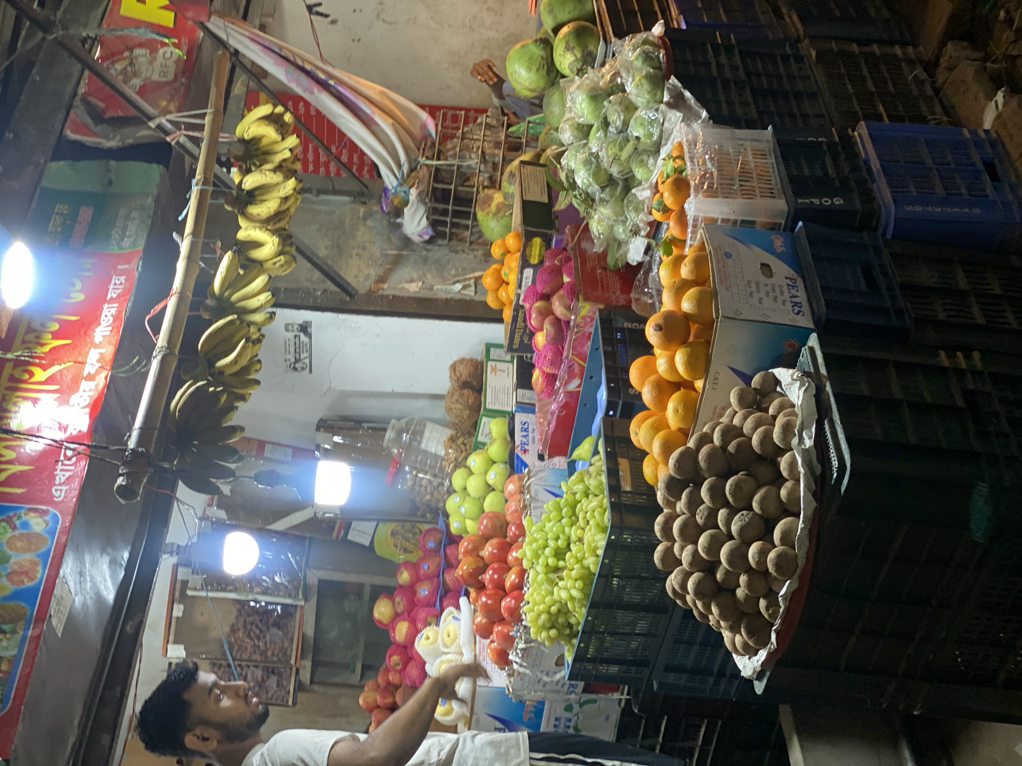 Here's a concise description of the image:
The image depicts a fruit stand in Dhaka, Bangladesh, at night. A man stands near the stand, seemingly tending to it. The stand is densely packed with a variety of fruits, including bananas, coconuts, guavas, oranges, apples, grapes, pomegranates, and sapodilla. Fruits are displayed in plastic crates and boxes. Above the fruit display, there are string lights and a sign in Bengali indicating the availability of all kinds of fruits. The background shows urban elements such as buildings.