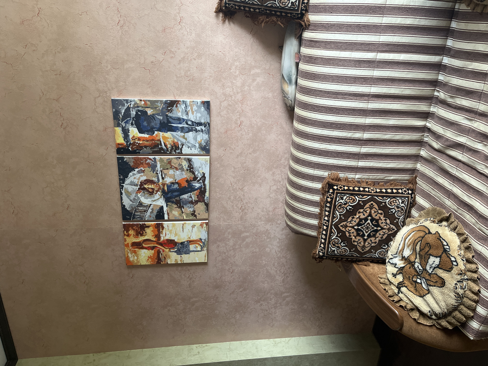 This is a medium shot of a living room interior in Vladimirovka, Russia. Three framed paintings depicting figures in a rainy cityscape hang on a textured, pinkish-brown wall. A striped sofa with brown and beige stripes is in the foreground, adorned with two decorative pillows. One is square with a patterned design, and the other is round with a depiction of a rabbit. A fish-shaped pillow is visible on the sofa behind the patterned pillows. The overall scene suggests a domestic setting.