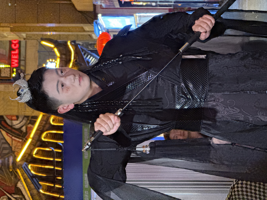 A young adult individual, appearing male, with fair skin and dark hair styled with shaved sides and a top ponytail secured by an ornate silver winged headpiece, stands in a dynamic pose at night in an urban area of Yuzhong District, China. The individual wears an elaborate black costume, featuring a patterned inner garment, a wide textured black belt/waistband, and flowing sheer outer sleeves with beaded tassels. Gold-colored embellishments are present below the outer corners of the eyes. Both hands grip a slender, metallic object resembling a sword or staff, held diagonally across the body. The individual's gaze is directed slightly off-camera to their left, with a neutral expression. In the blurry background, behind the main subject's left arm, the upper portion of another individual's face is partially visible. The environment is heavily lit by artificial illumination, including prominent yellow/orange neon lights on a background building that mimic traditional Chinese rooflines, a partial red neon sign reading "WELCO," a red Chinese lantern, and various other blue and indistinct commercial signs, indicating a vibrant street or market setting.