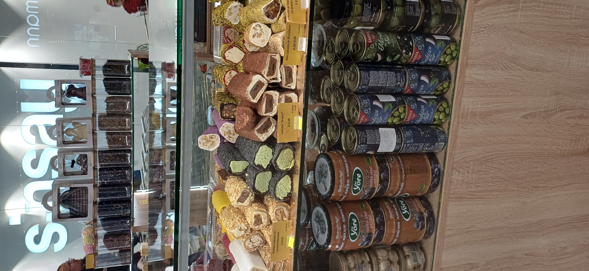 The image shows a display of Turkish delight and canned olives in a store. In the foreground, there are several rows of Turkish delight pieces, some coated in pistachios, others in chocolate or nuts. Small yellow price tags are visible in front of some of the Turkish delight assortments, indicating prices in Euros. Below the Turkish delight, there are several large brown cans labeled "Yöre" with images of olives, and stacked next to them are smaller, blue and white cans of olives, some with Spanish text. In the background, the store's sign "sinzai" is visible in large white letters on a blue background. To the left, a man wearing glasses is partially visible, looking towards the display. The lighting suggests an indoor environment, likely a food shop or market.